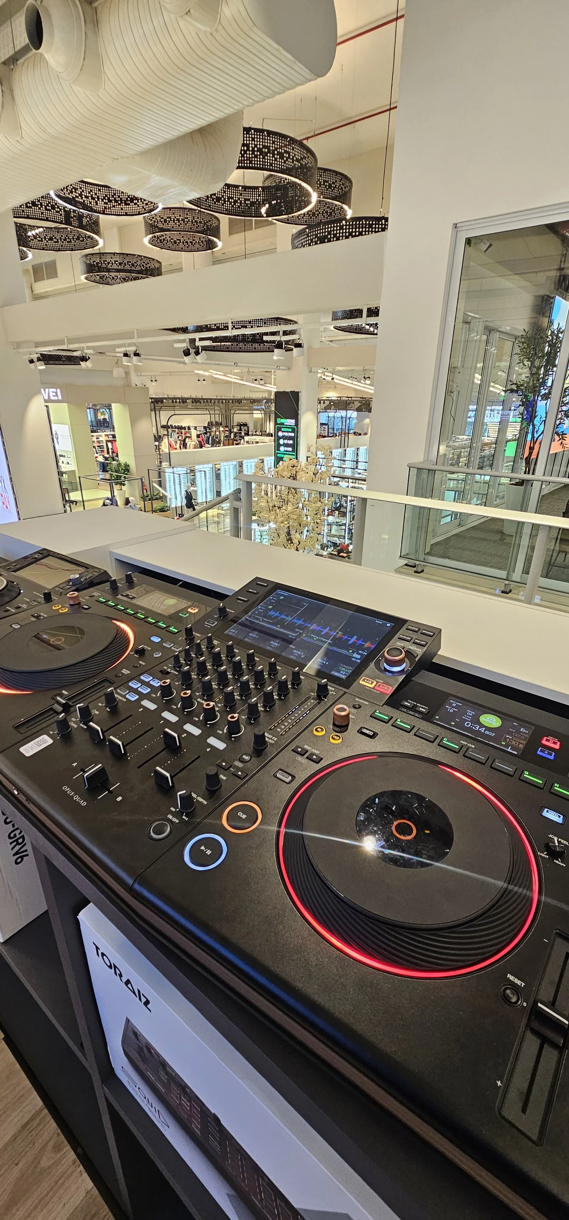 Close-up of a professional DJ mixing console with a shopping mall interior in the background, including clothing stores and decorative ceiling lights.