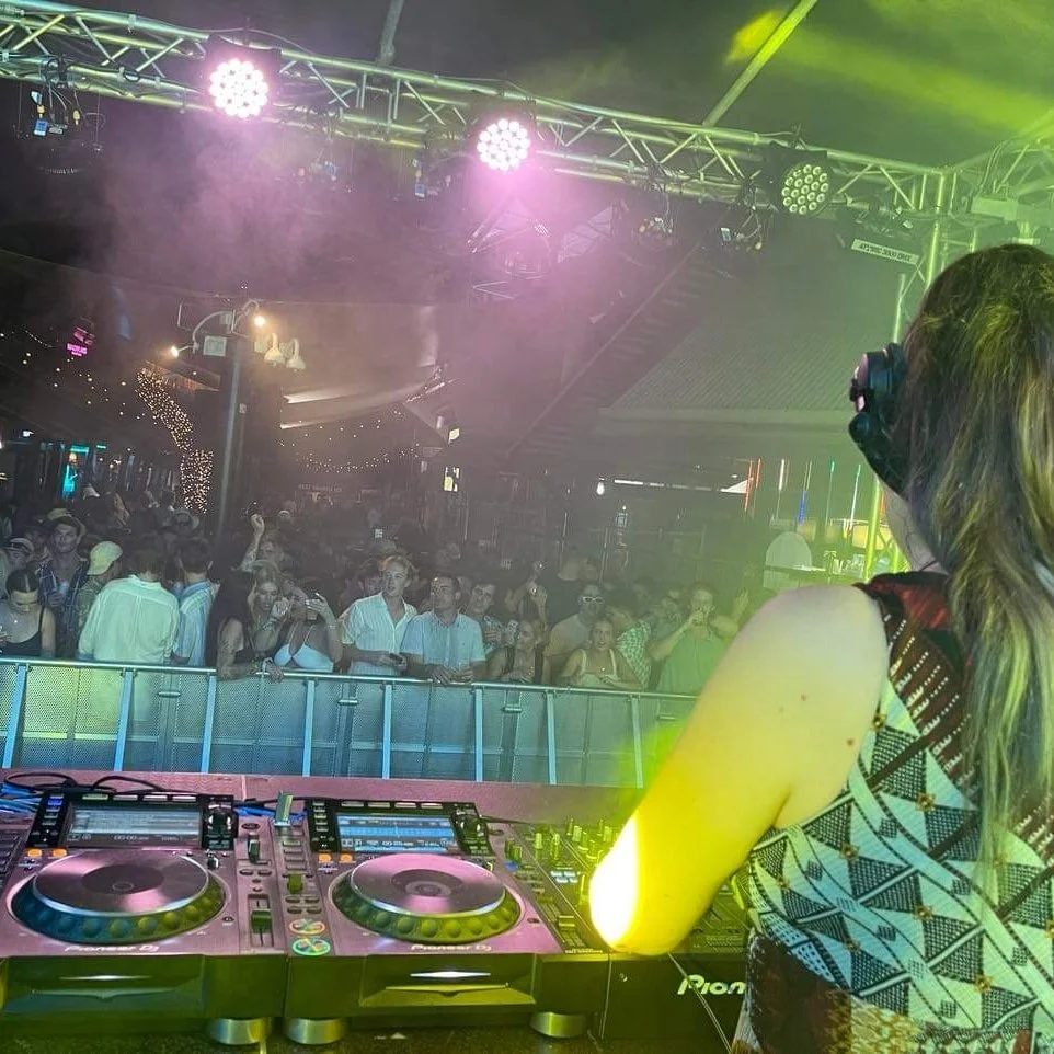 A DJ performing at a club or concert with a crowd dancing in front of her. The scene features colorful stage lighting and fog effects, creating an energetic atmosphere.