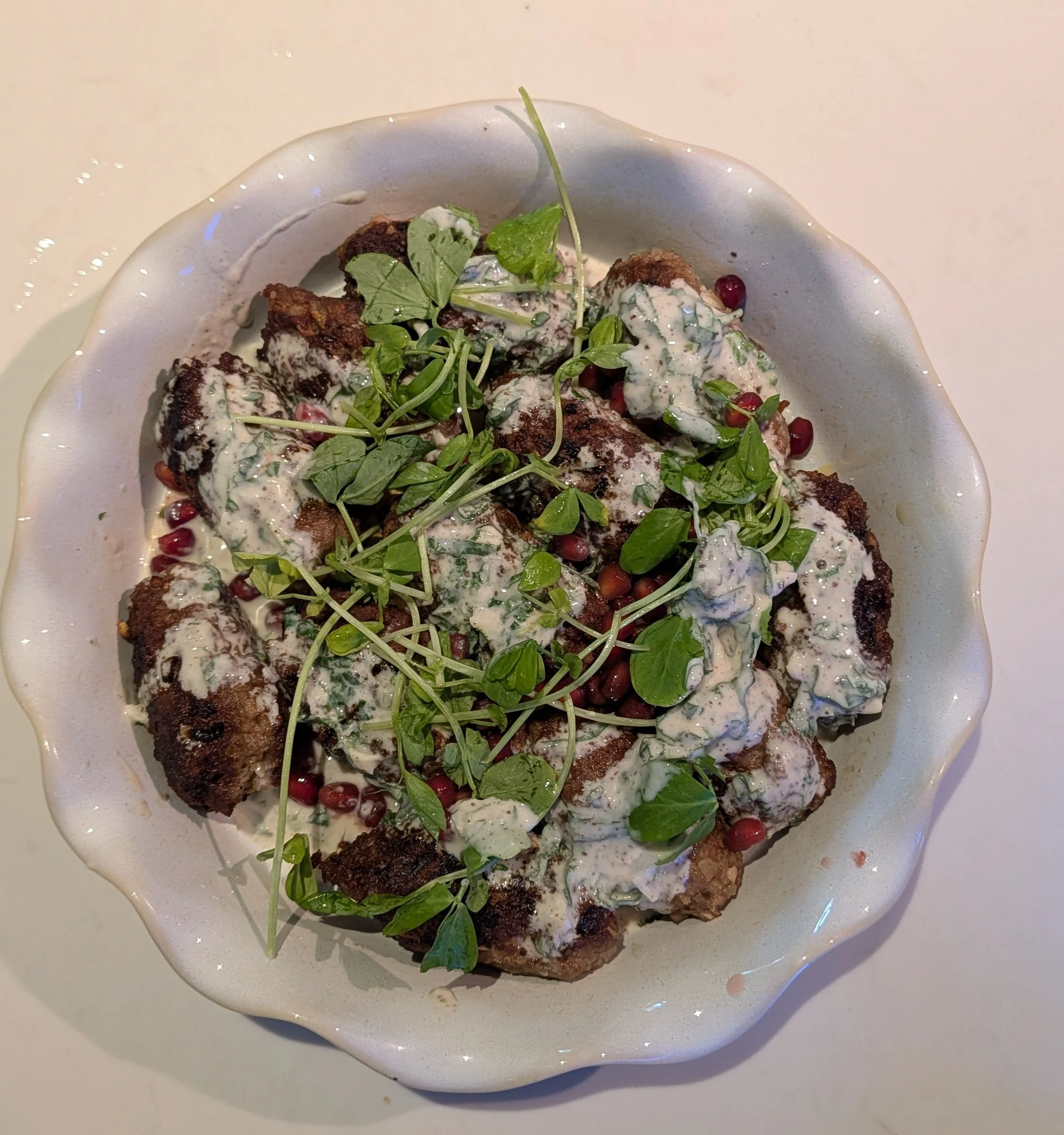 Plate of lamb kofta topped with green herbs and drizzled with white sauce, garnished with pomegranate seeds and green microgreens.