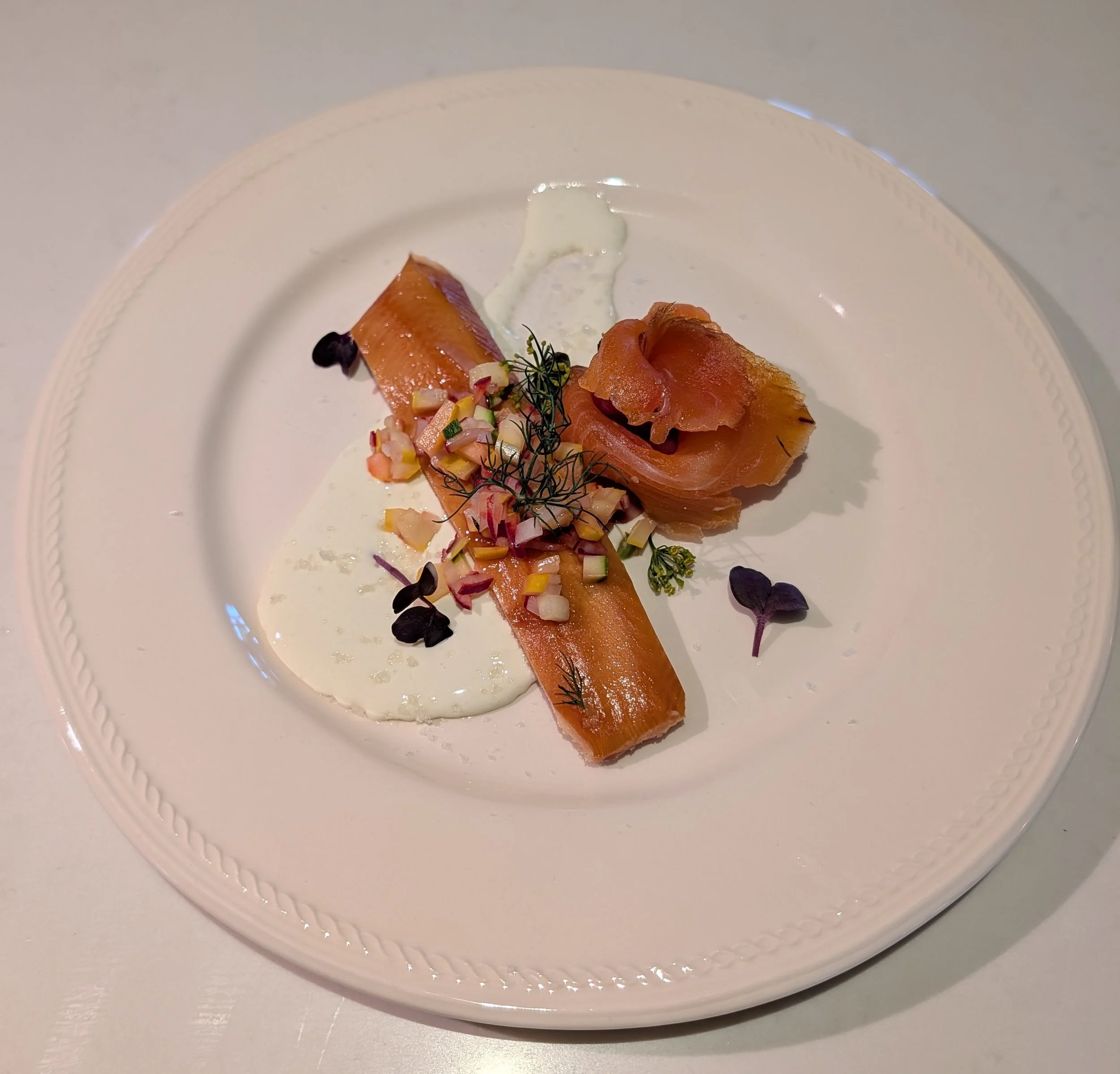A plated dish featuring smoked salmon, garnished with chopped vegetables and microgreens, accompanied by a creamy white sauce and small dark edible flowers.