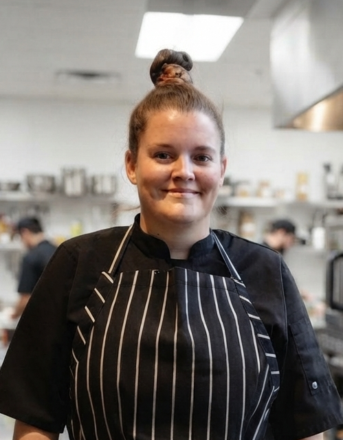 A professional chef in a black uniform and striped apron smiling warmly in a commercial kitchen.