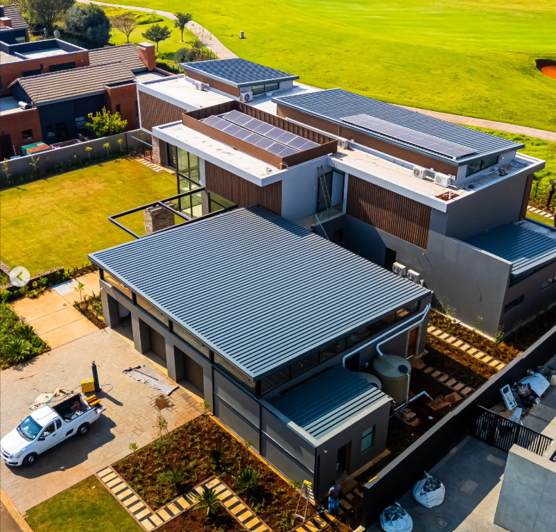 An aerial view of a modern residential house with a metal roof, solar panels, and adjacent lawn and garden areas.