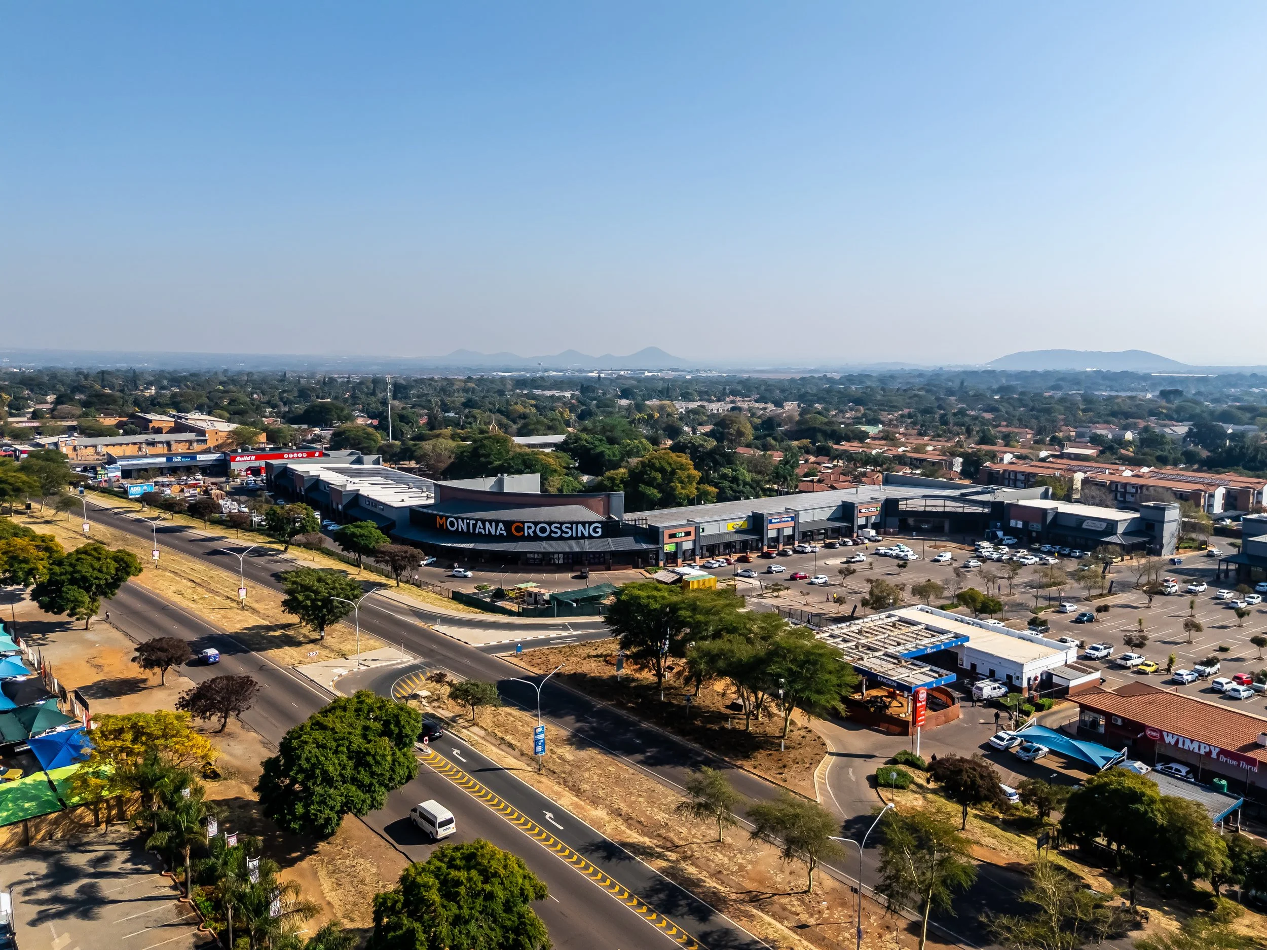 Aerial view of a commercial area with a shopping center named Montana Crossing, parking lot, and surrounding residential neighborhood under a clear blue sky.