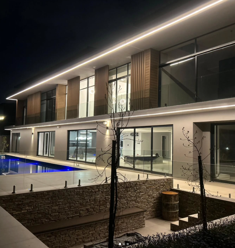 Night view of a modern multi-story house with large glass windows, illuminated exterior lighting, a swimming pool, and a stone retaining wall with small trees in the foreground.