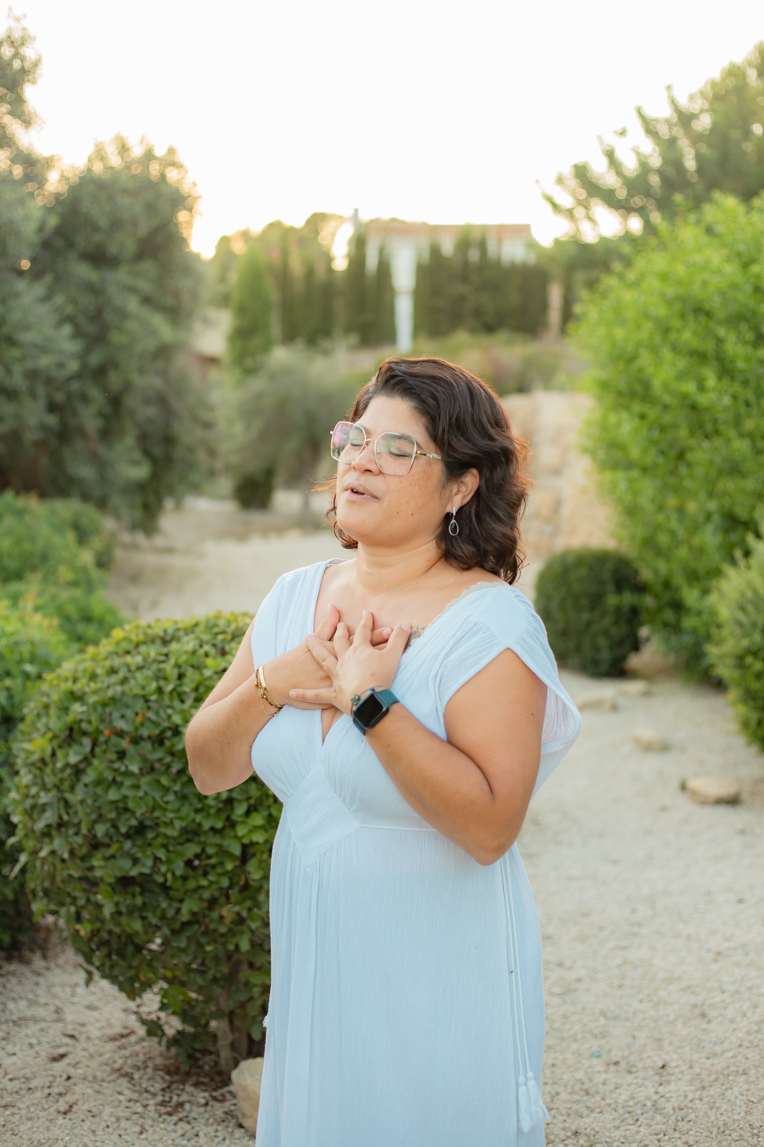 A woman with glasses and short brown hair in a light blue dress standing outdoors with her hands on her chest, eyes closed, appearing to be in a moment of prayer or reflection, surrounded by greenery and sunlight.