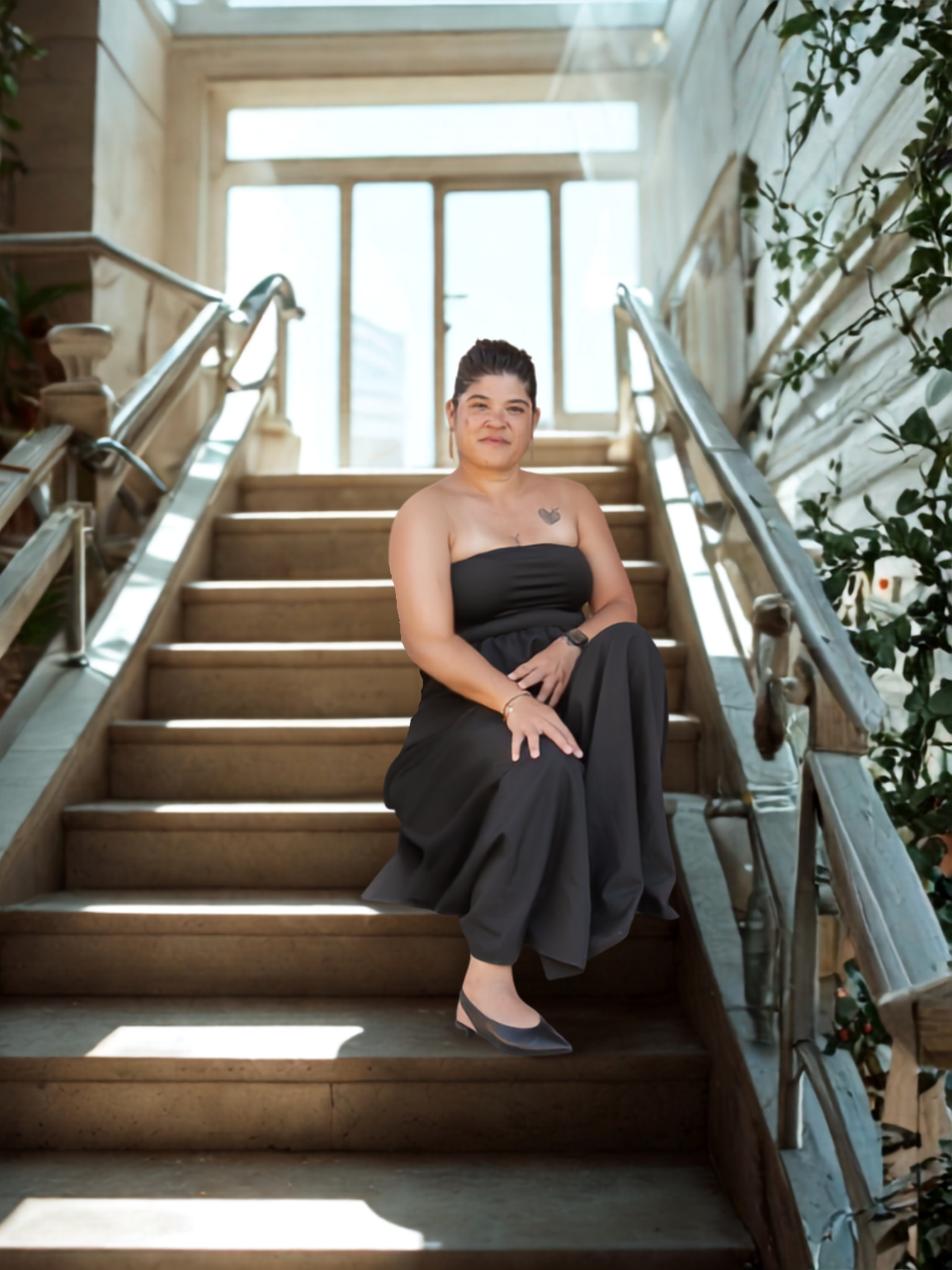 A woman sitting on wooden stairs near a large window, wearing a black strapless dress and heels, with plants on the side.