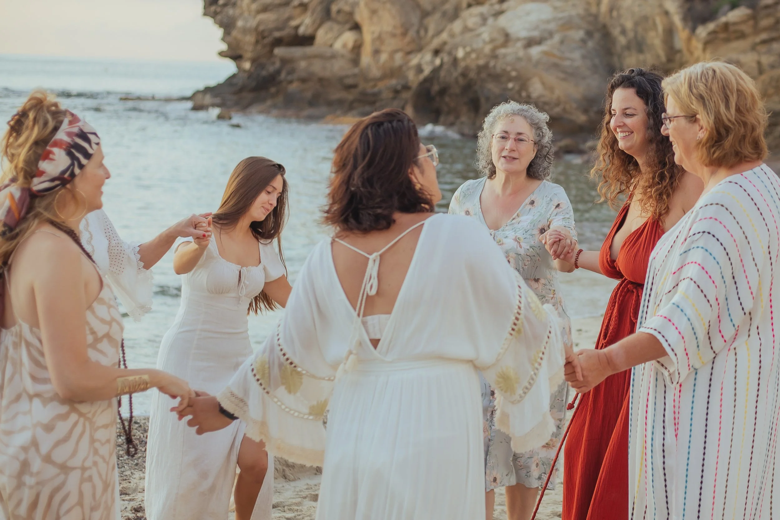 A group of women holding hands in a circle on a beach near rocks and water, smiling and engaged in a moment of connection.