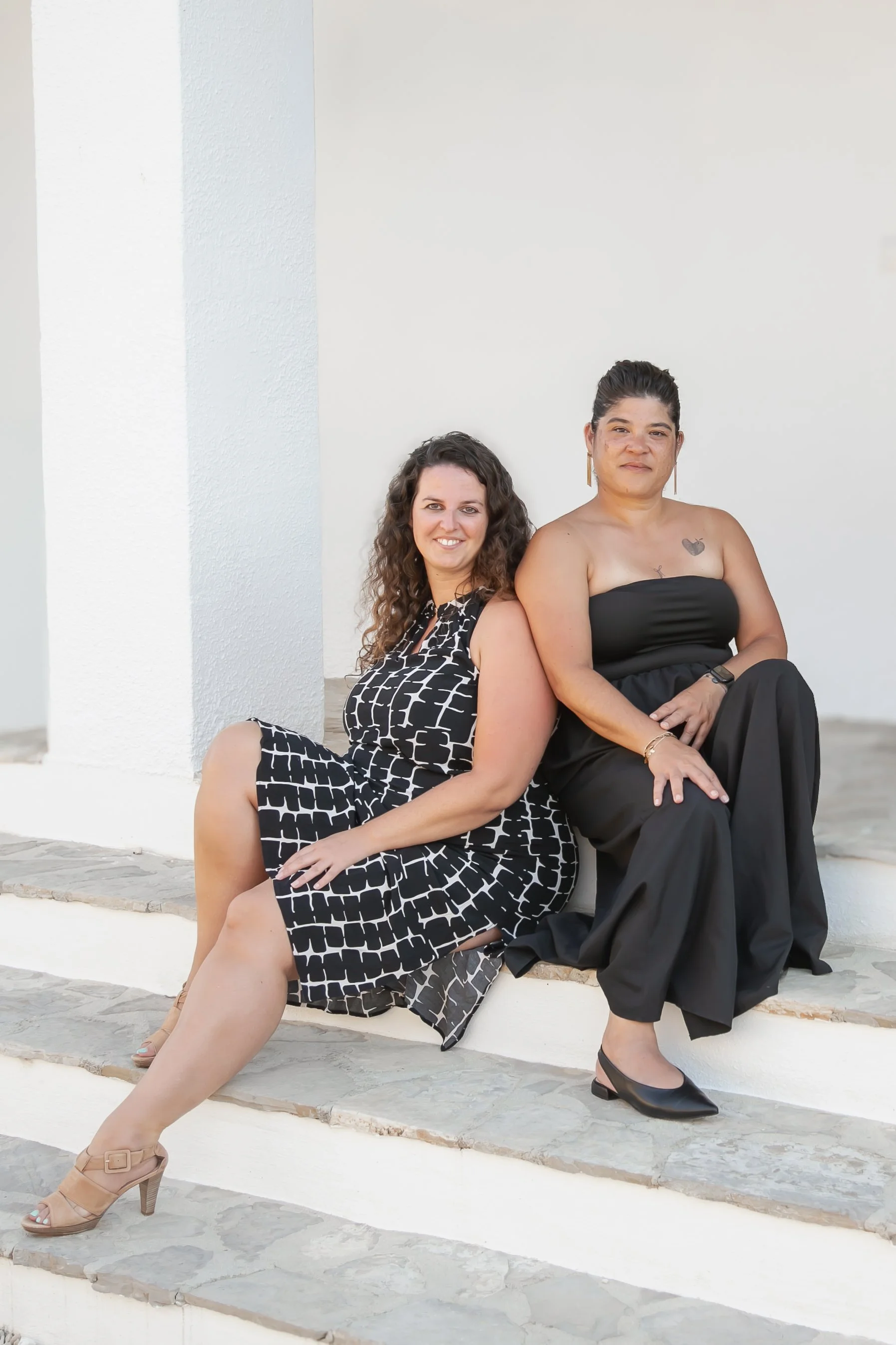 Two women sitting on white steps against a white wall, smiling at the camera. One woman has long curly hair, wearing a black and white patterned dress and beige heels. The other woman has dark hair pulled back, wearing a strapless black jumpsuit and black heels.