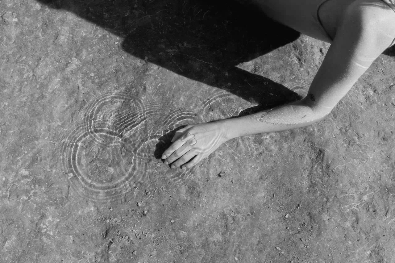 A person's elongated arm and hand reach out to touch the ground, creating ripples in a small puddle on rough, textured earth surface in black and white.