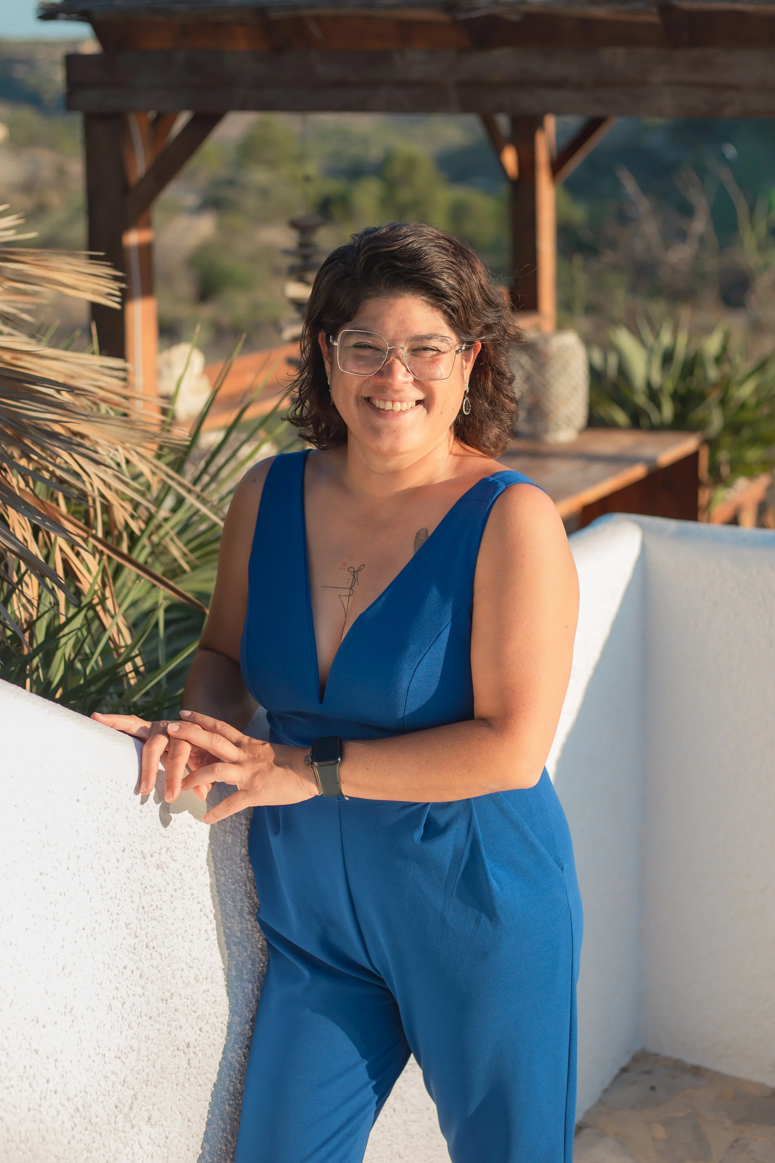A woman wearing glasses and a blue dress, smiling and standing outdoors by a white wall with greenery and wooden structure in the background.