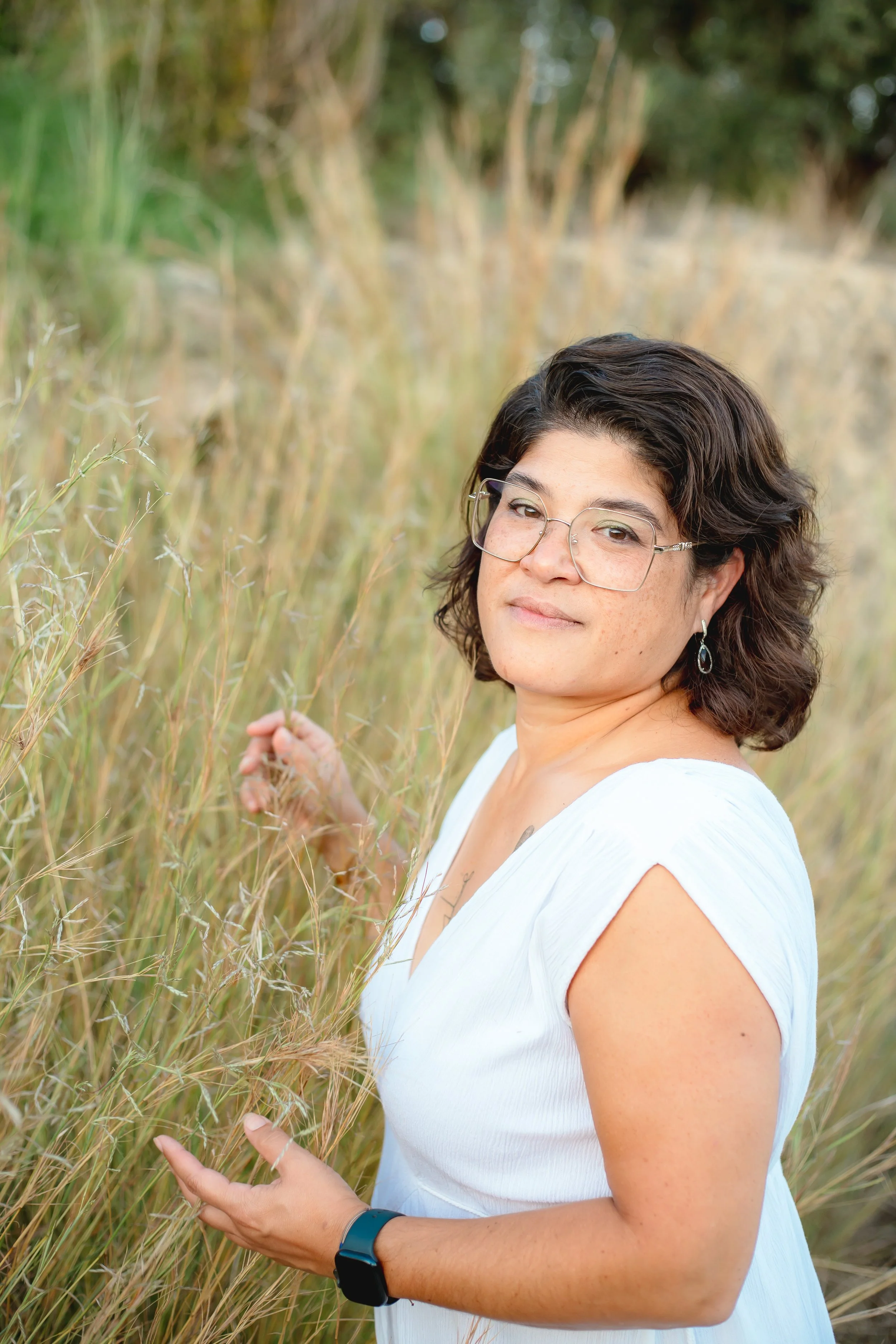 A woman with dark wavy hair and glasses standing in a field of tall, dry grass, with trees in the background.