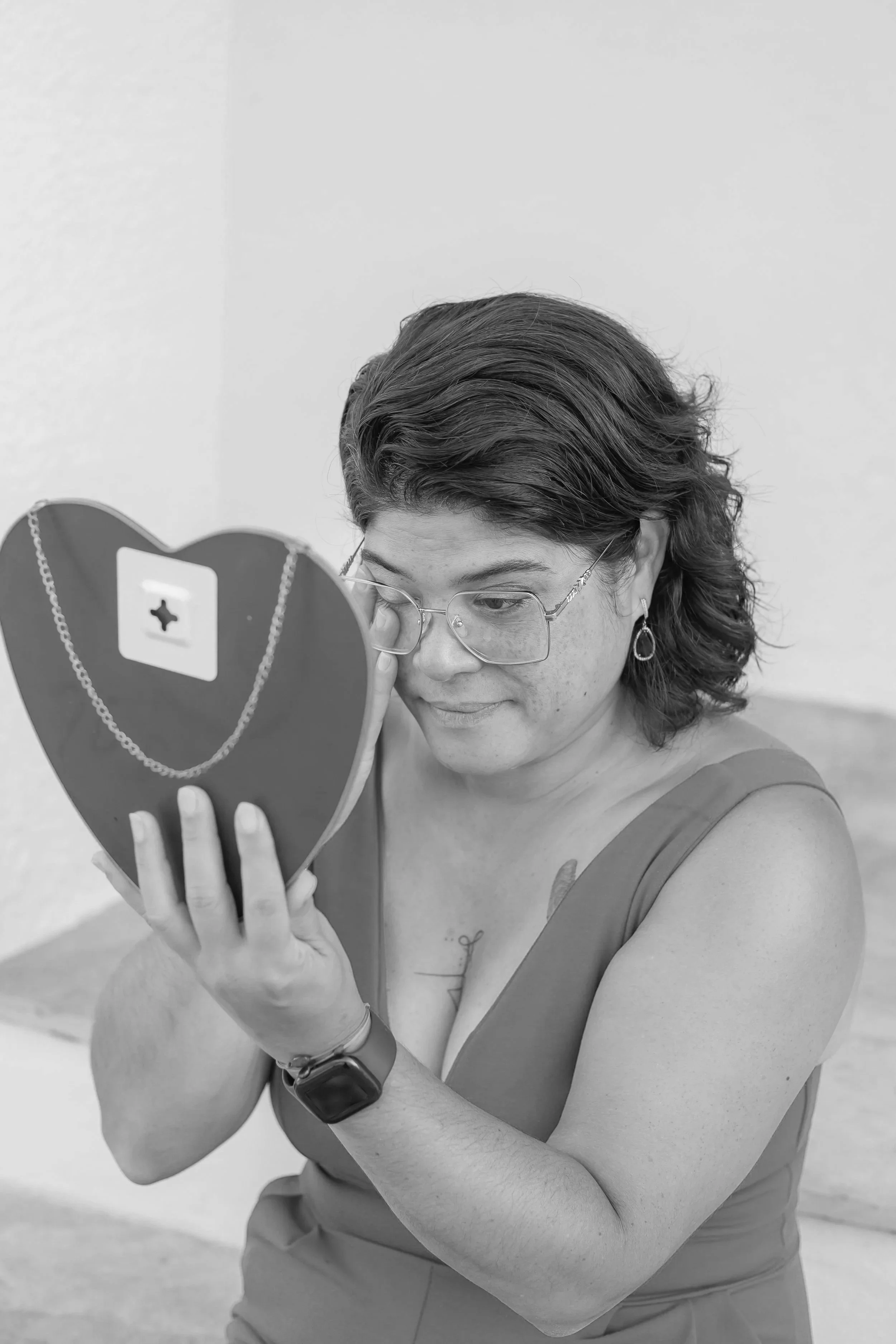 A woman with curly hair and glasses holding a heart-shaped mirror, looking at her reflection.