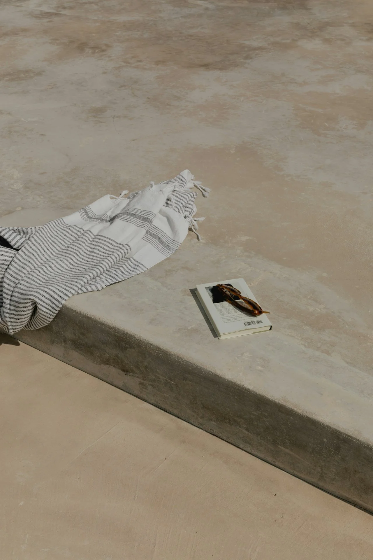 A striped fabric cloth partially covers a beige stone bench, with a white book and a pair of sunglasses placed on top of the book, on a stone floor.