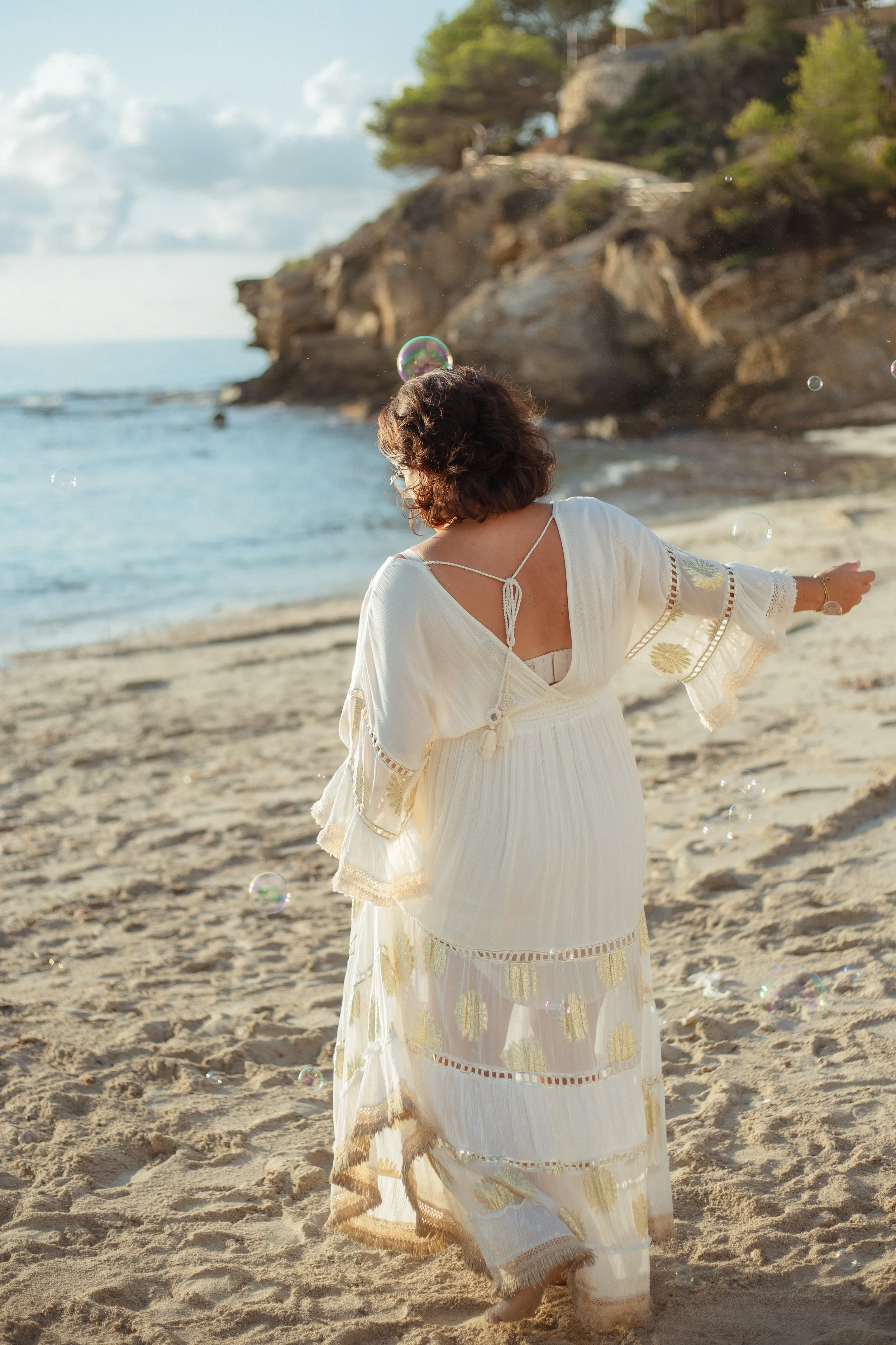 A woman in a white dress standing on a sandy beach with her back to the camera, surrounded by soap bubbles, with rocky cliffs and trees in the background.