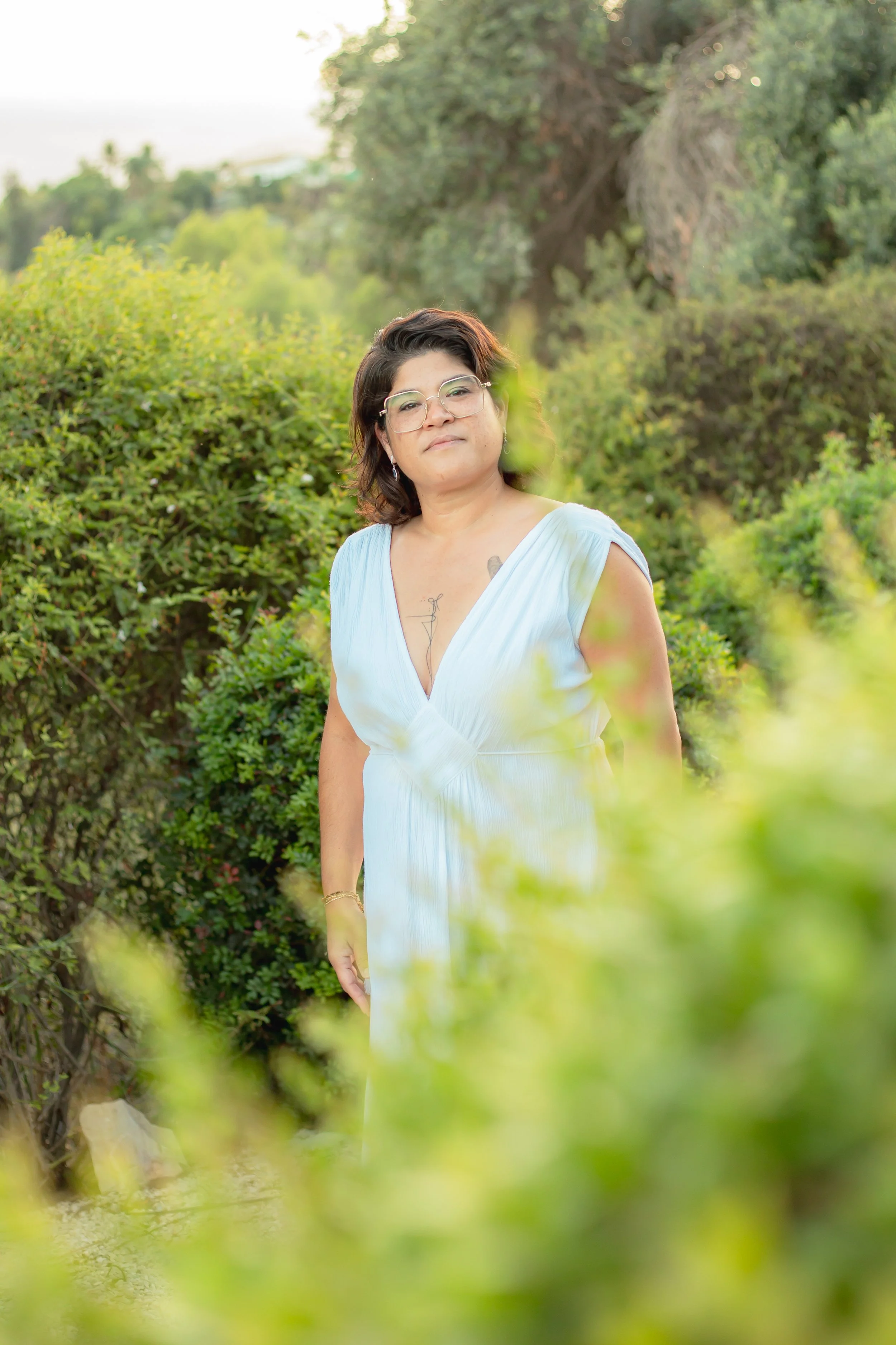 A woman with short dark hair, glasses, and earrings standing outdoors among lush green bushes and trees, wearing a light blue dress. The background shows hills and a partly cloudy sky.