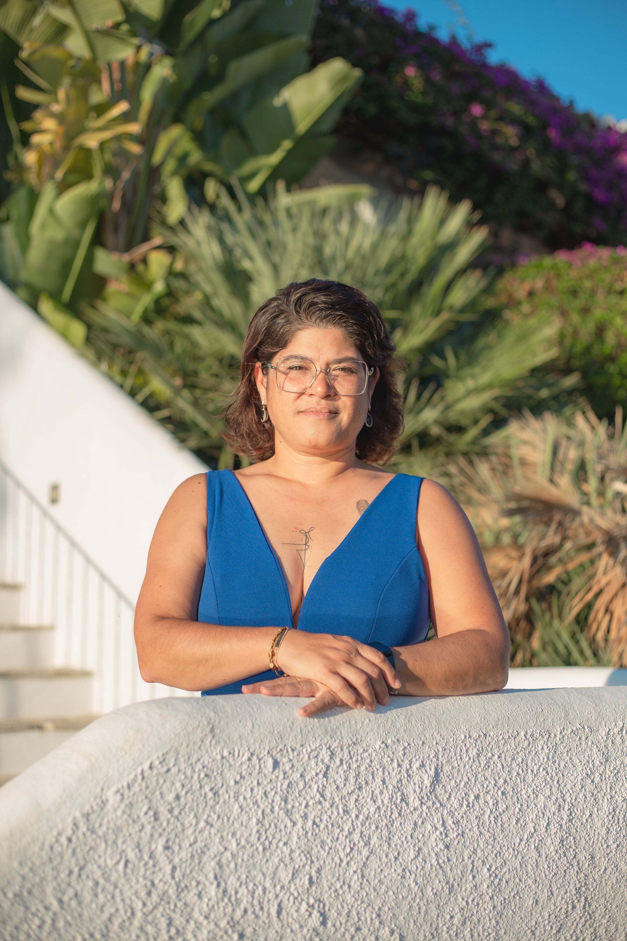 A woman with glasses wearing a blue dress, standing outdoors with a background of large green plants and purple flowers, leaning on a white curved surface