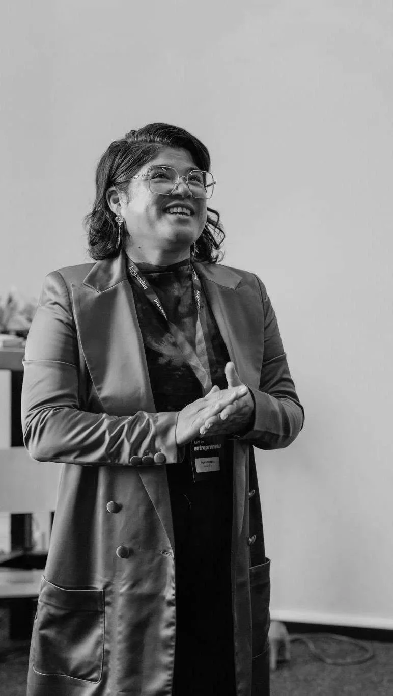 A woman with shoulder-length hair, glasses, and earrings standing and smiling in a professional setting, wearing a blazer, shirt, and a conference badge around her neck.