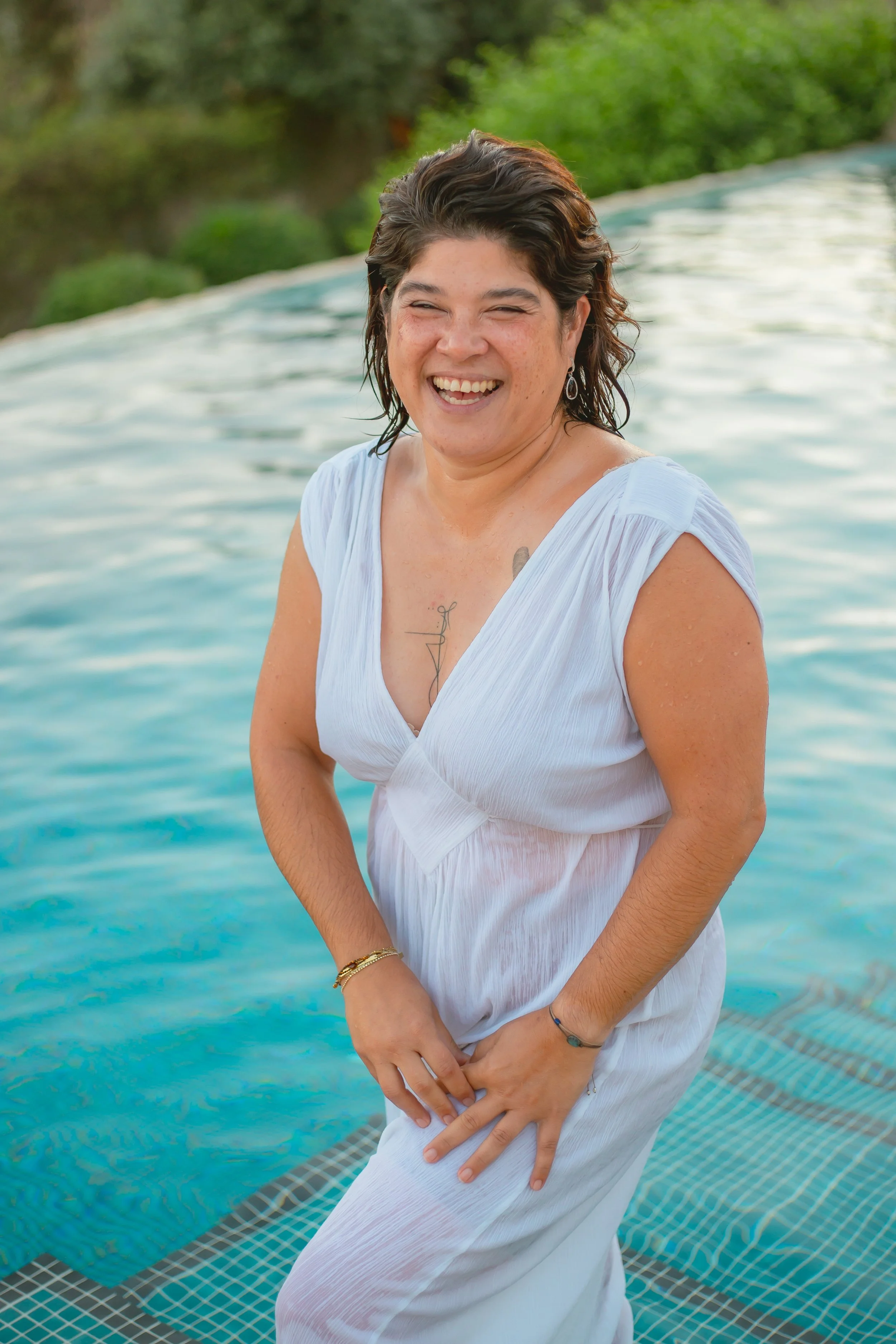 Woman standing in a pool, smiling, wearing a white dress with water droplets, greenery in the background.