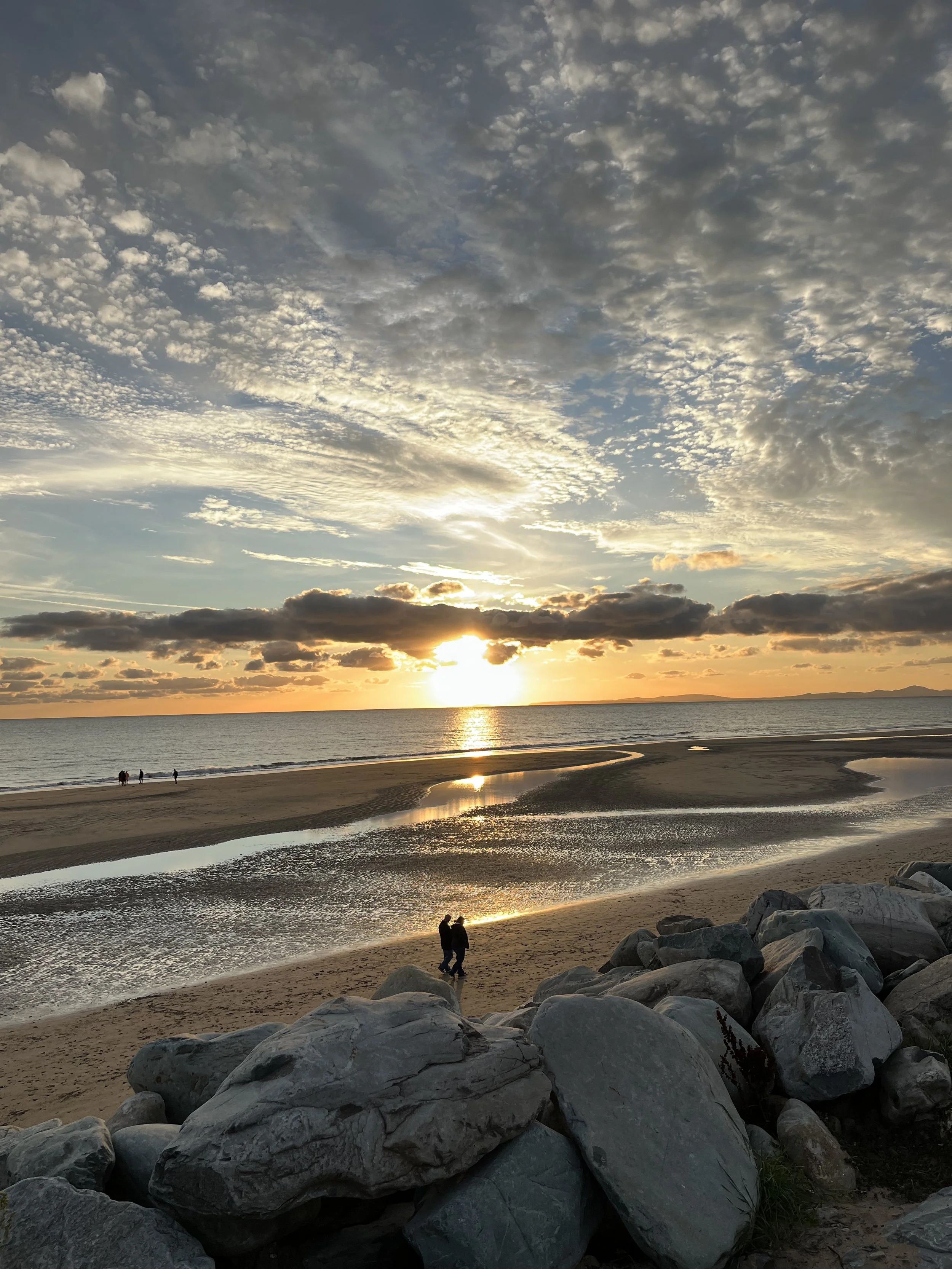 A beach at sunset with the sun near the horizon, casting a golden glow over the sky and reflecting on the water. There are a few clouds scattered across the sky, and several people are walking along the shoreline.