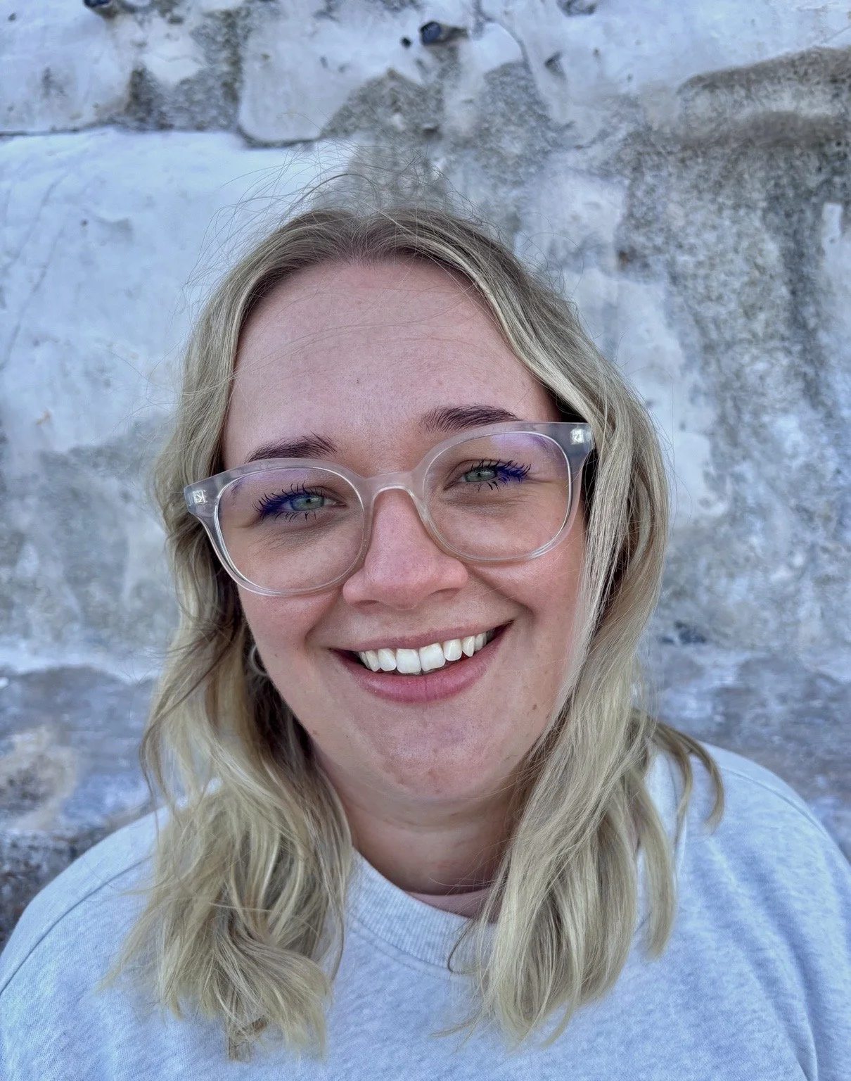 A woman with blonde hair and clear glasses smiling in front of a textured stone wall.