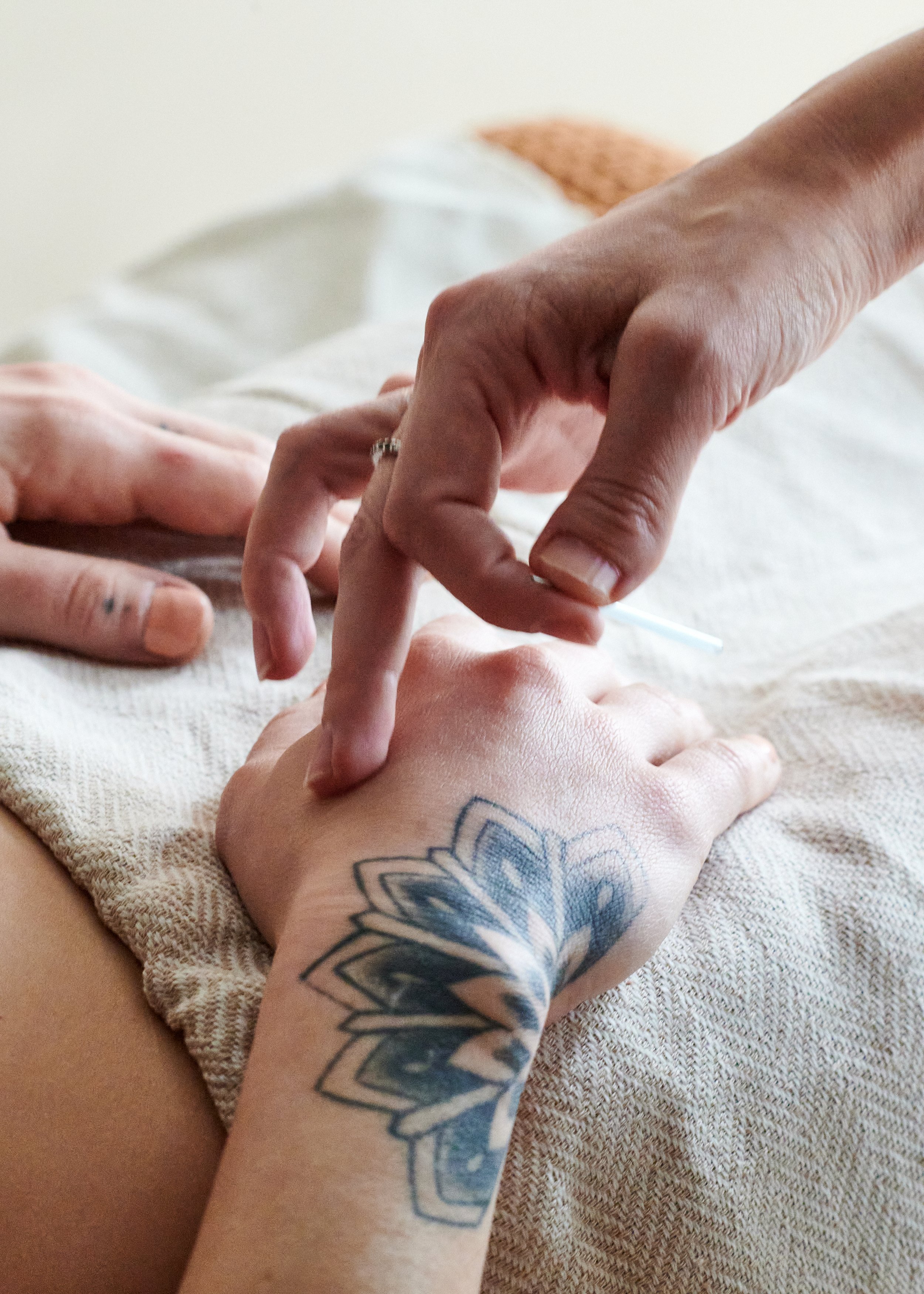 Close-up of someone receiving a tattoo on their arm, with the tattoo artist's hand in focus. The person's arm has a colorful flower tattoo, and the scene appears to be in a tattoo studio.