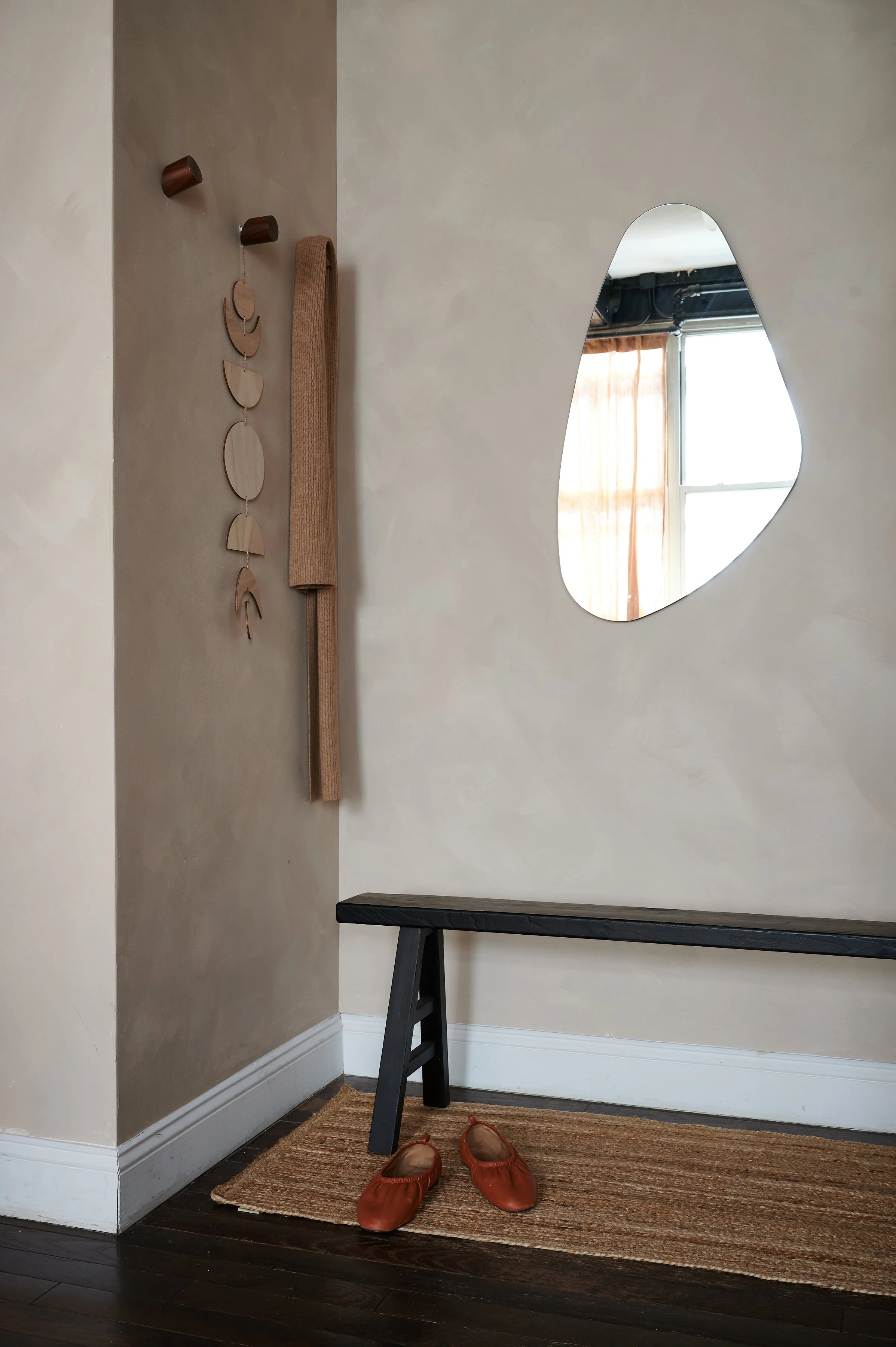 Entryway with a mirror, hooks with a brown blanket and wall hanging, a black bench, a pair of brown slippers, a woven rug on dark wood floor, beige and brown walls, and a window with curtains.