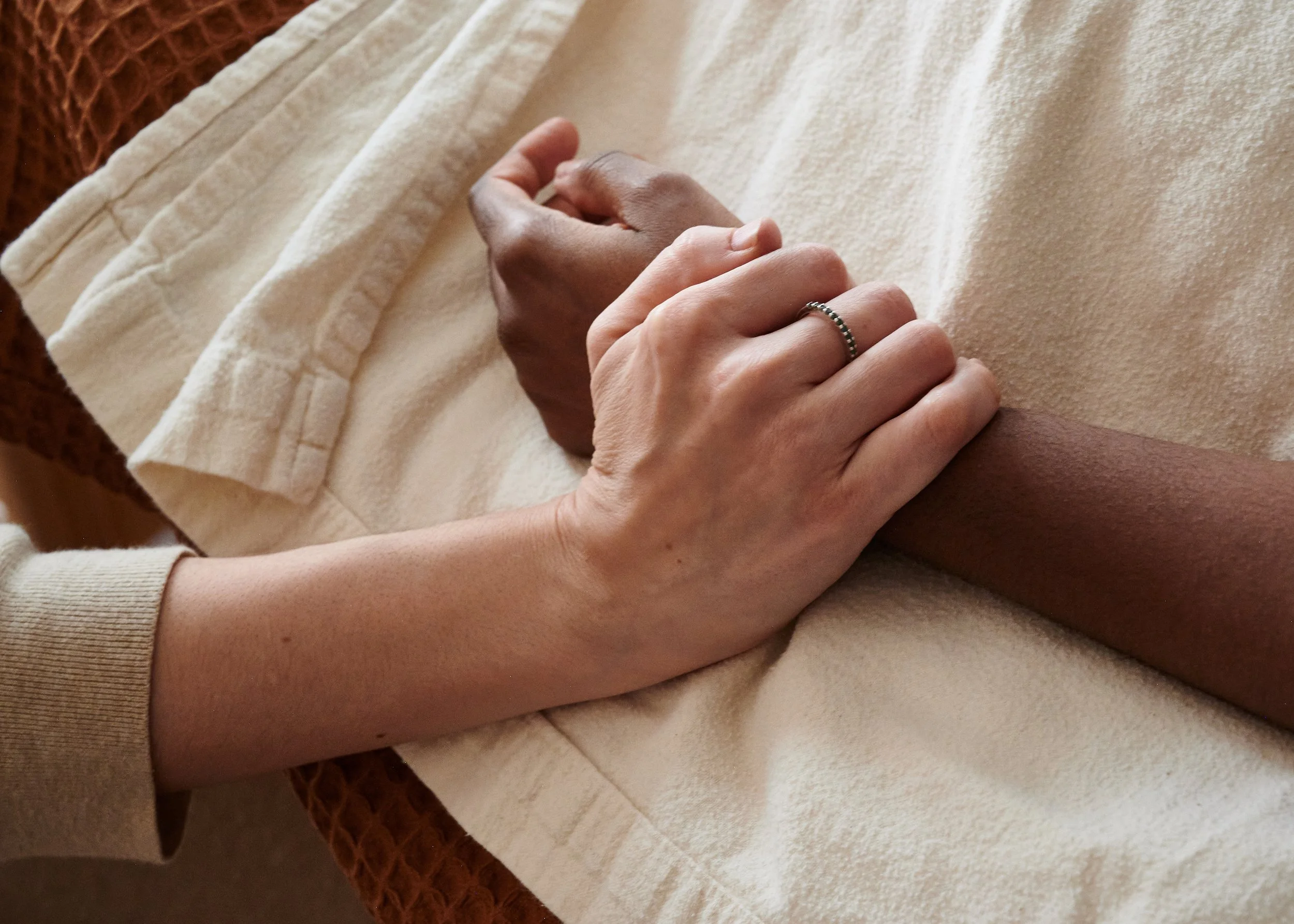 Close-up of two people holding hands, one with a ring, resting on a cream-colored blanket.