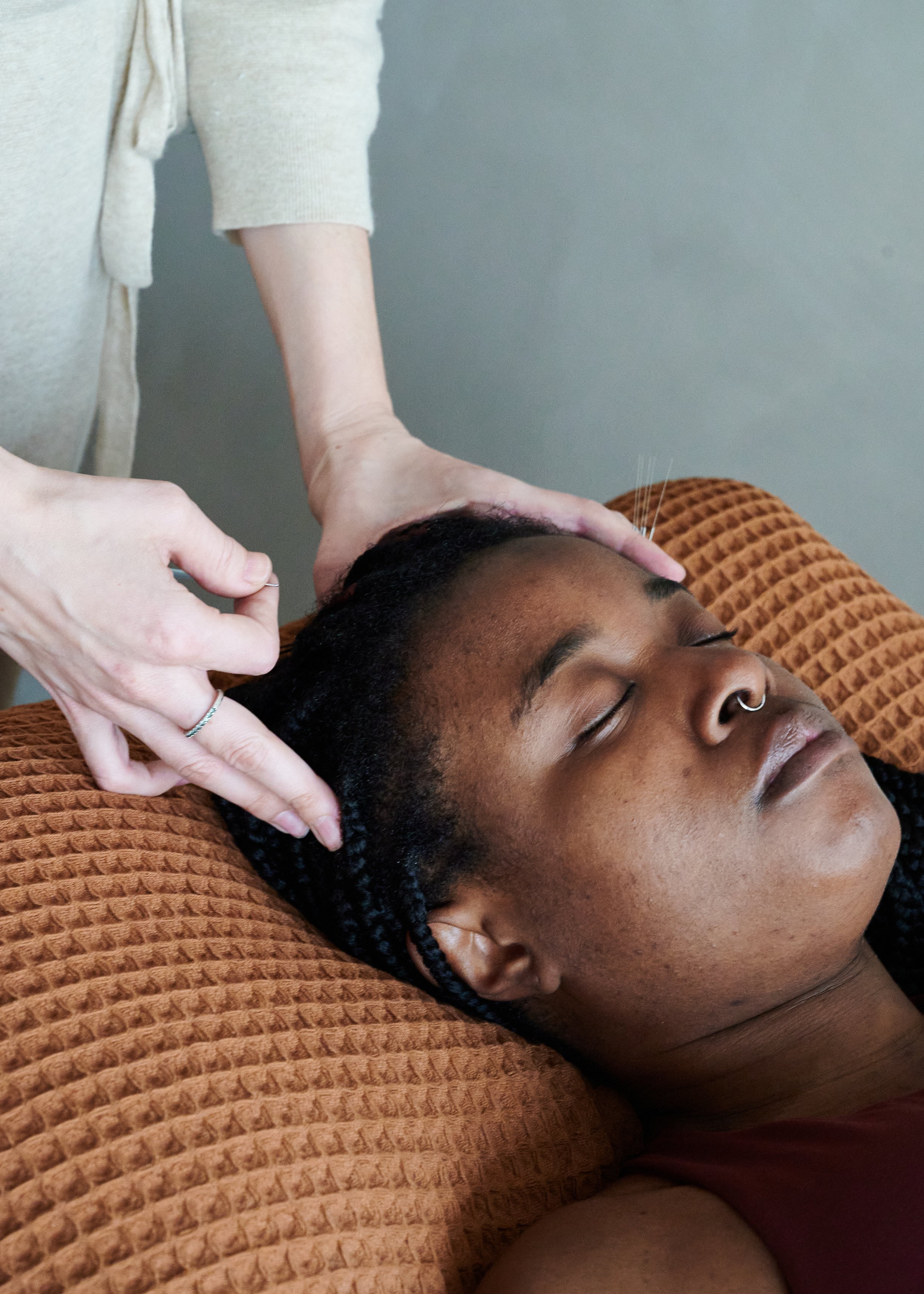 A person with dark braided hair receiving a head massage while lying down on a textured, orange blanket.