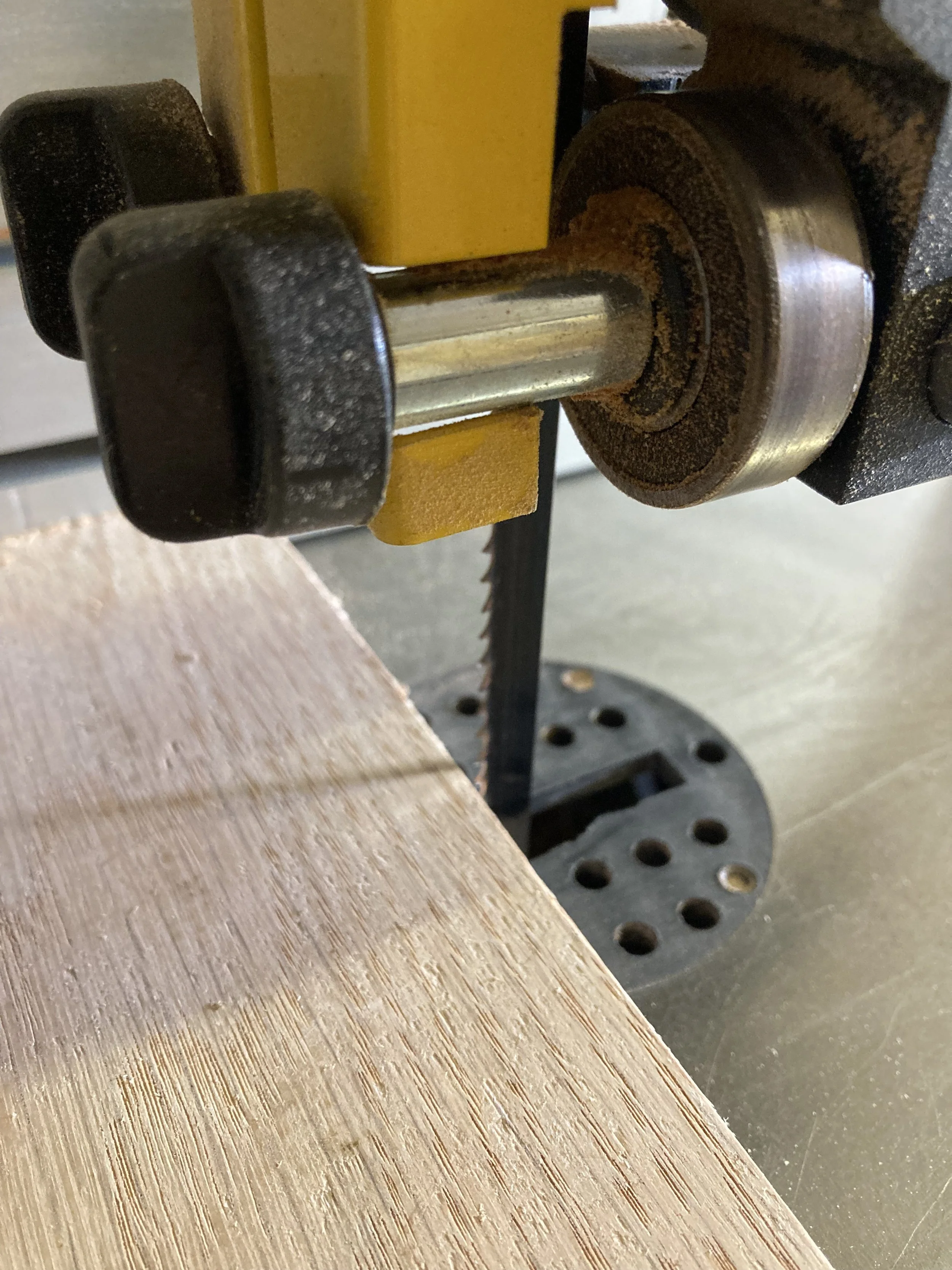 Close-up of a woodworking bandsaw blade with a yellow guide roller and black tension knob, cutting a piece of wood with visible grain.