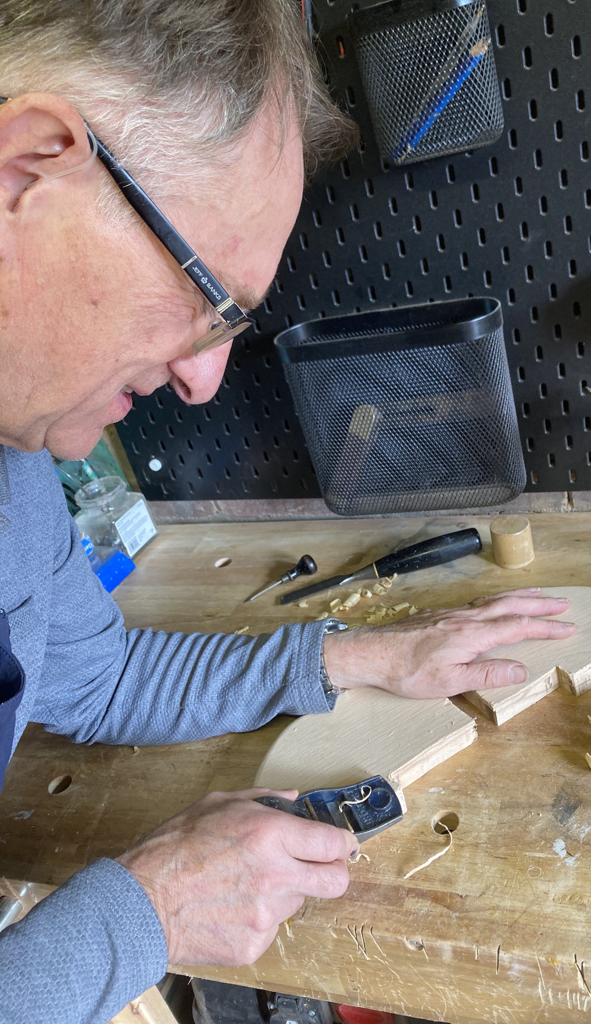A man wearing glasses is woodworking at a workbench, using a power drill and carving a piece of wood. The workspace has various tools and wood shavings, with a black pegboard holding a pen and pencil in the background.