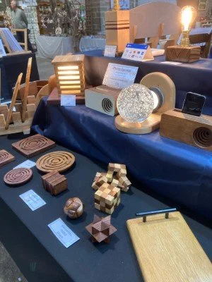 Display of wooden and cardboard home decor items, including lamps, coasters, and decorative sculptures on a table covered with dark fabric at an indoor market or craft fair.
