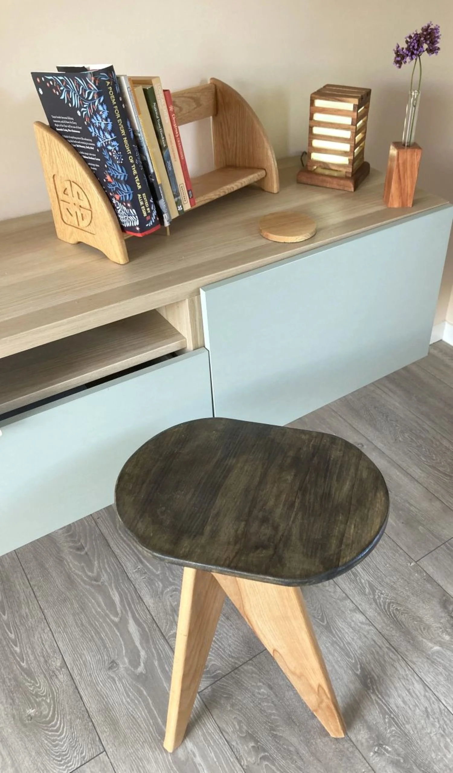 A wooden chair with a dark stained seat and light wooden legs, placed in a room with wooden flooring. In the background, there's a light wood cabinet, a small shelf with books, a wooden lamp, and a glass vase with purple flowers.