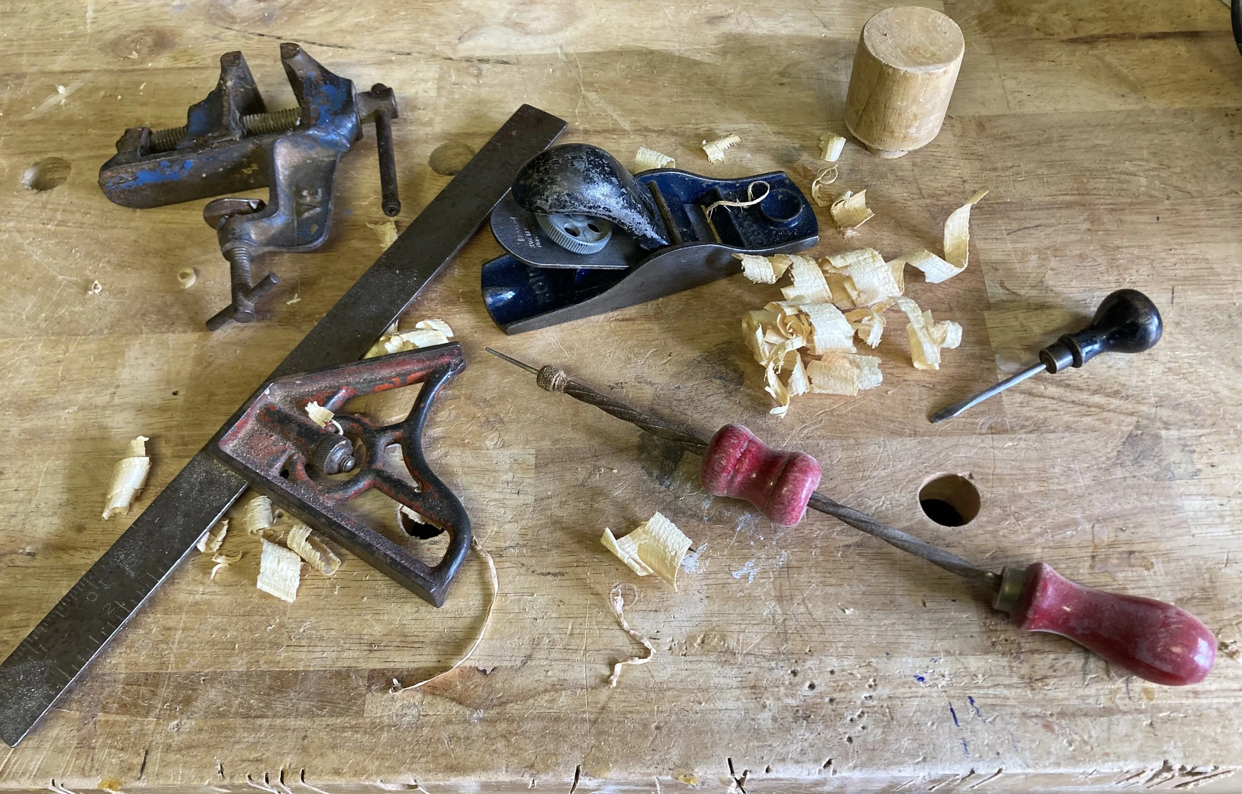 Woodworking tools on a workbench, including a hand plane, a saw, a chisel, and wood shavings.