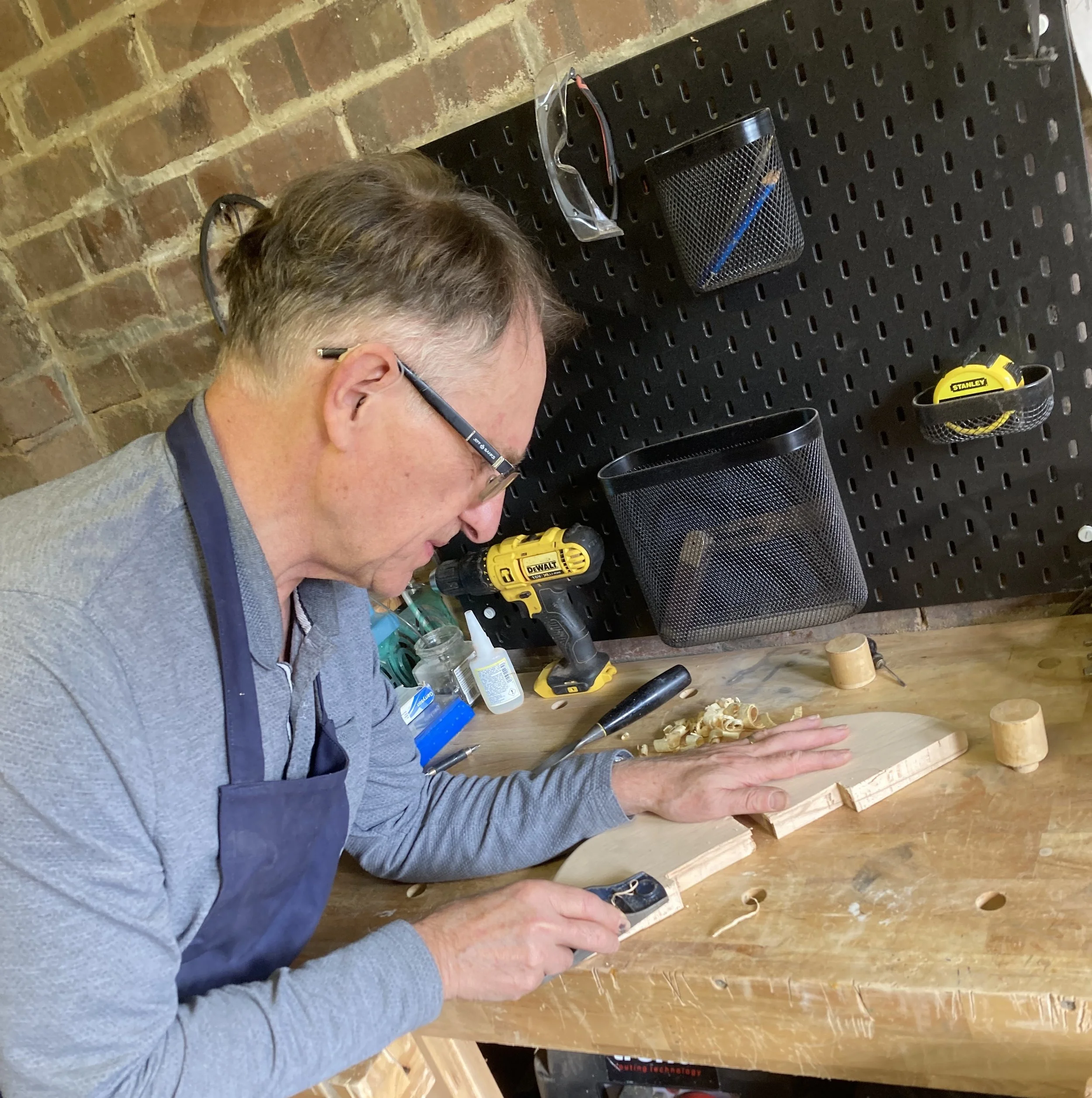 A person working with wood in a workshop with tools and materials on a workbench, using a cordless drill.