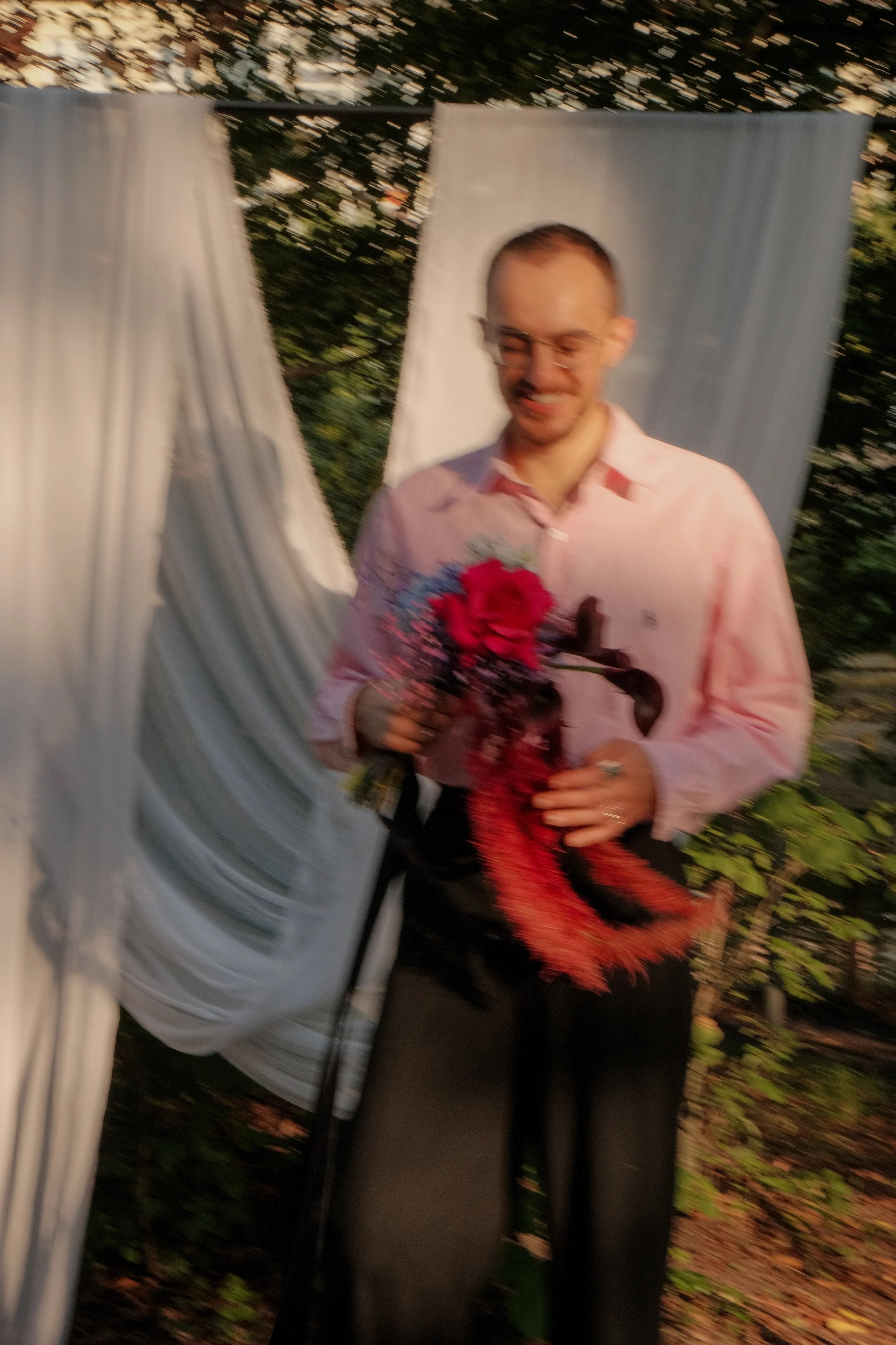 Blurred outdoor photo of a smiling man in glasses and a pink shirt holding a bouquet of red roses beside a person in a light blue shirt.