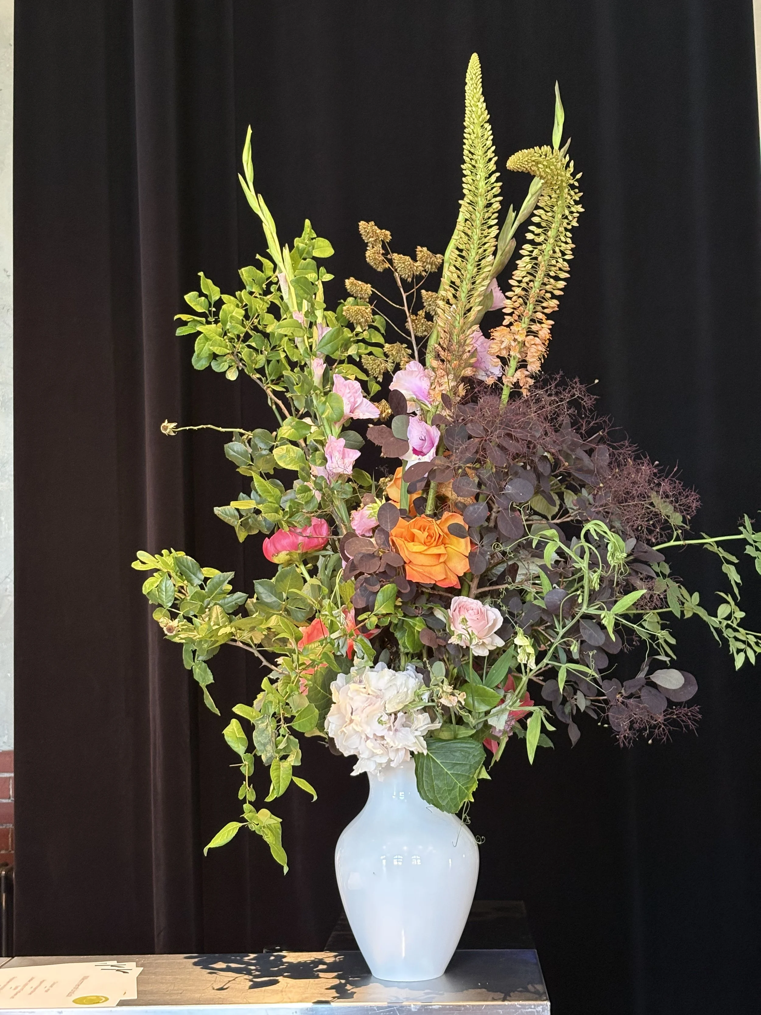 Large colorful wildflower arrangement with roses, greenery, grasses, and seed pods in a white vase against a black background