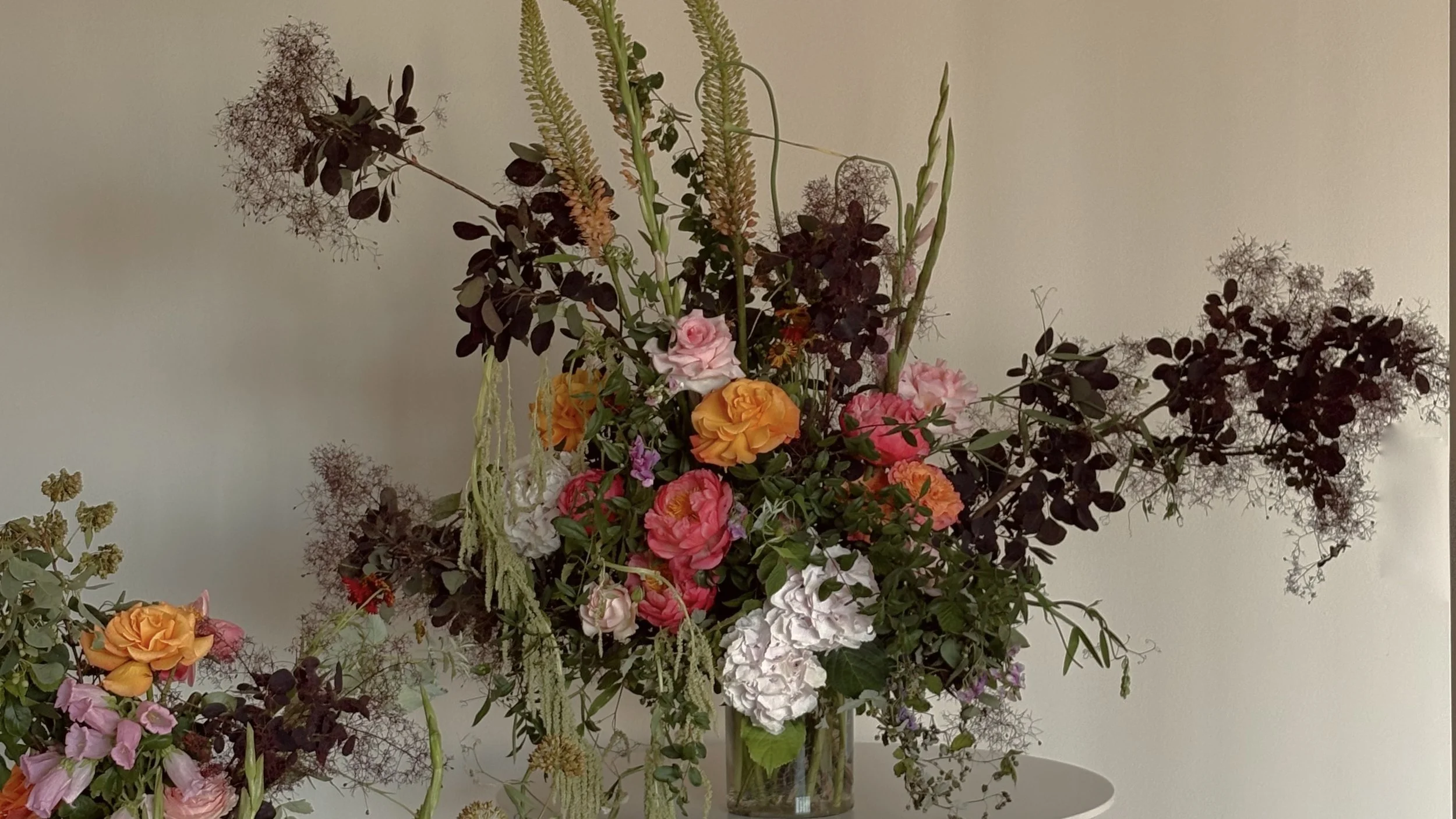 Large colorful floral arrangement with pink, orange, red, and white roses, greenery, and cascading branches in a glass vase