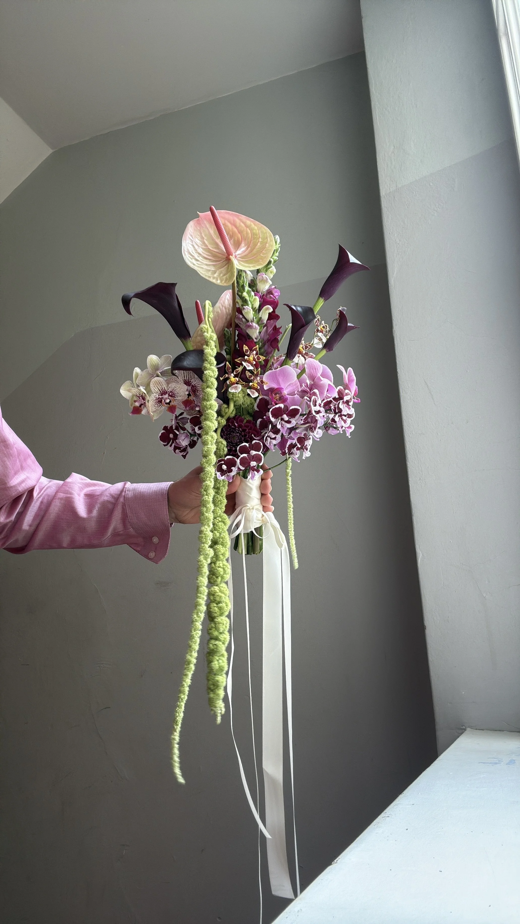 Hand holding an elaborate bouquet of anthuriums, calla lilies, orchids, and cascading green flowers tied with white ribbons.