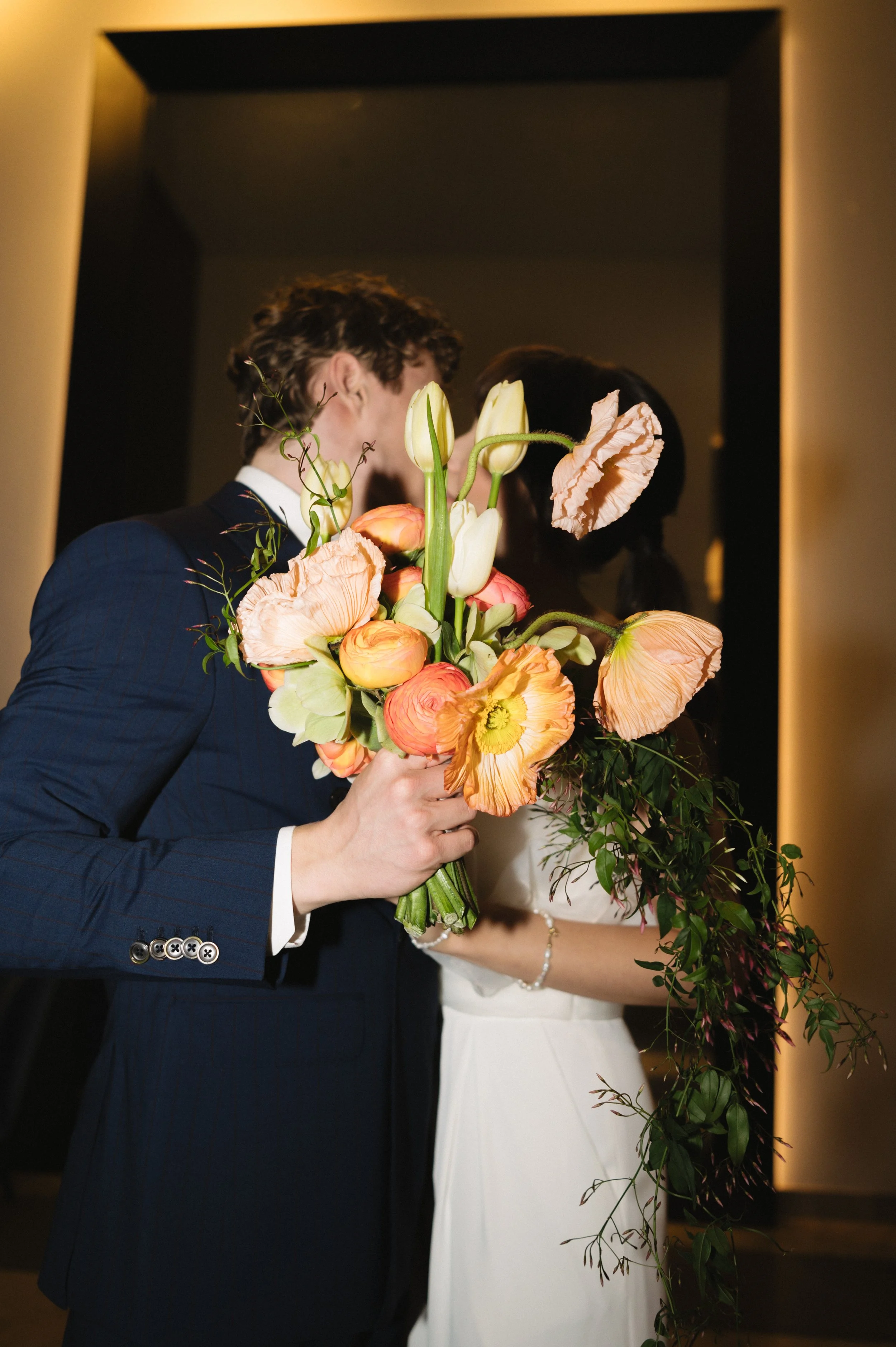 Couple in formal attire holding a lush peach and coral flower bouquet in a warmly lit room