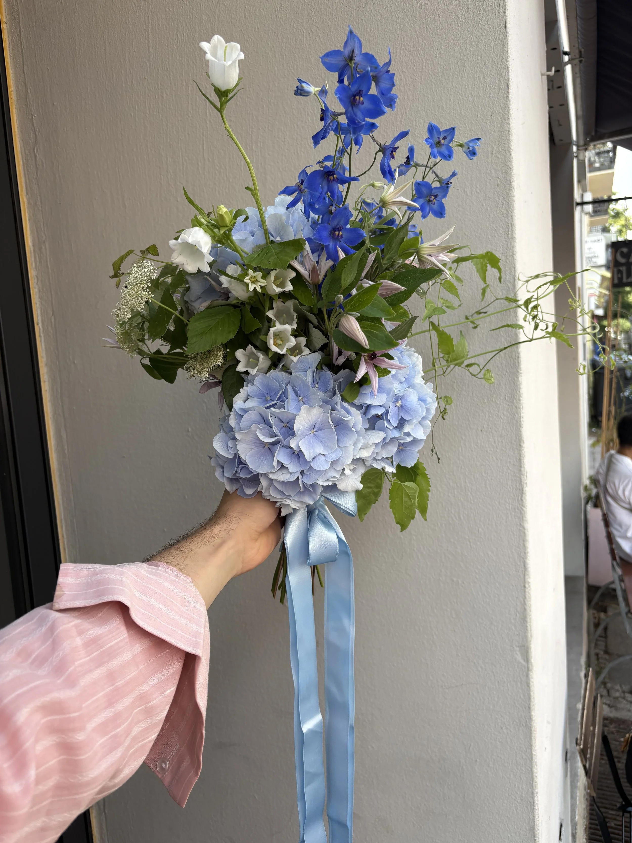 Hand holding a lush bouquet of blue hydrangeas, blue delphiniums, white flowers, pink lilies, greenery, and a light blue ribbon.