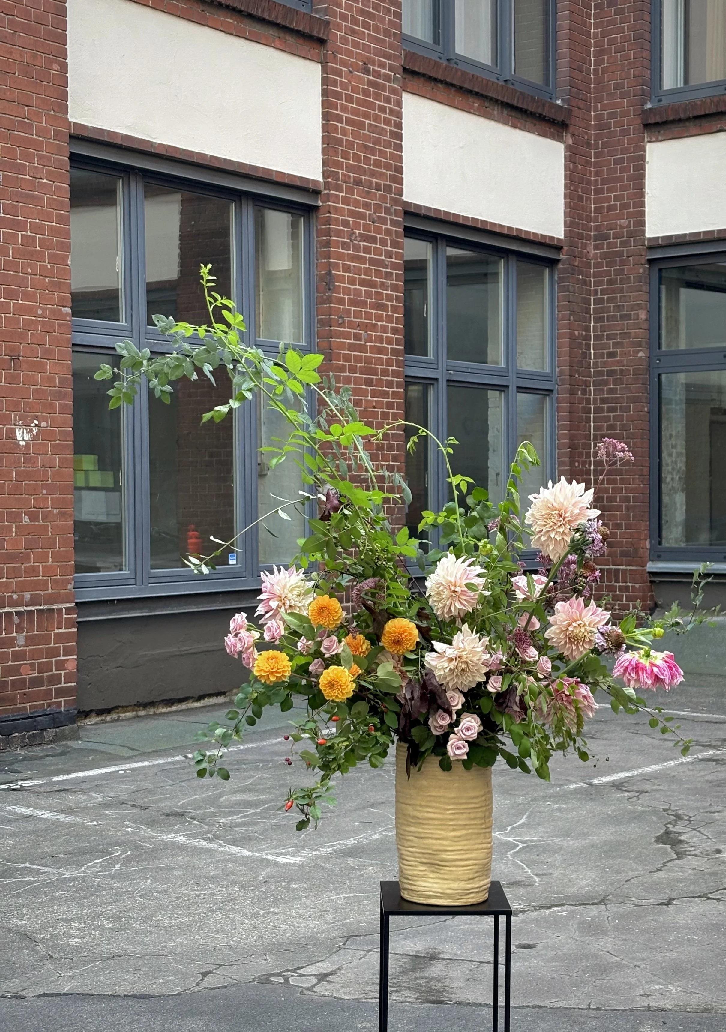 Large colorful flower arrangement in a textured vase on a black stand outside a red brick building