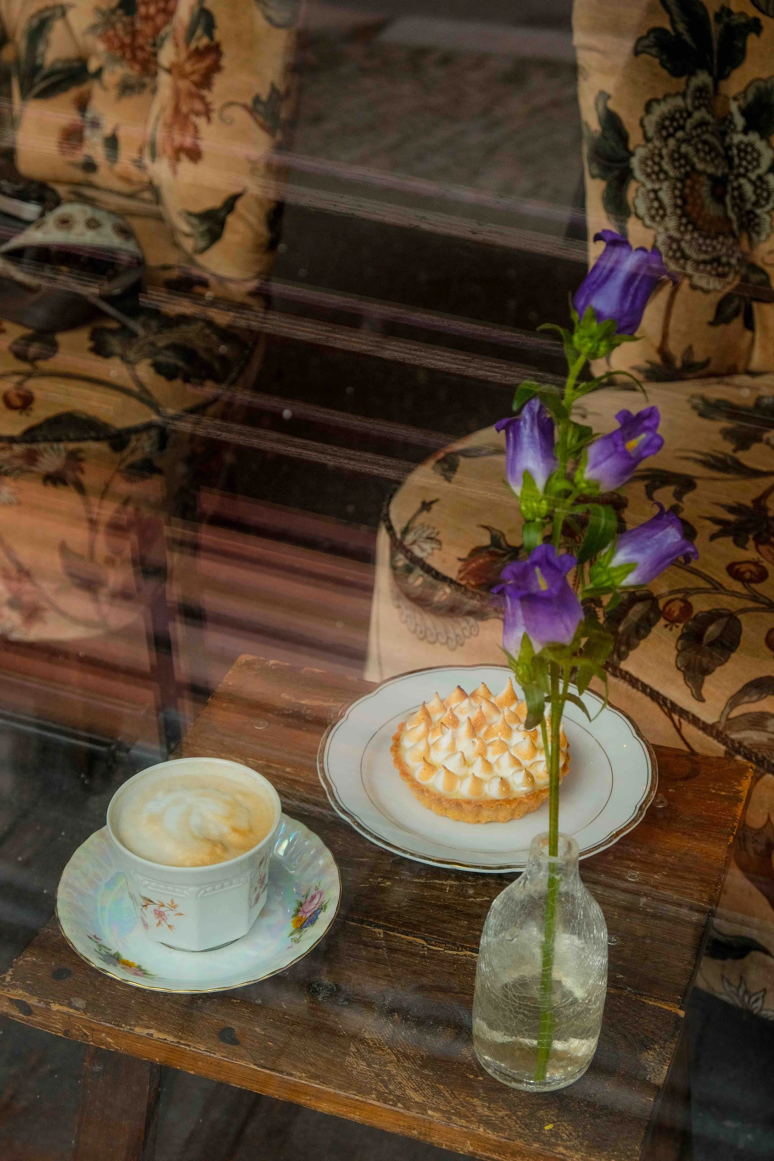 Vintage tea setting with a floral teacup, toasted meringue tart, purple flowers in a glass vase, and wooden shutters.