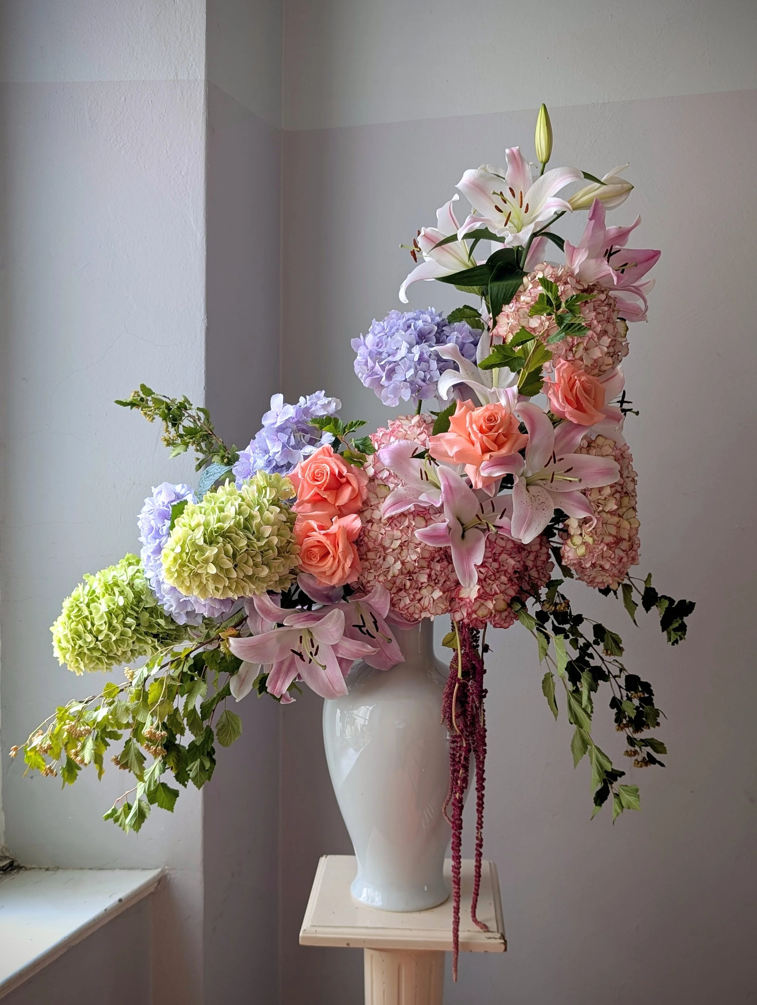 Abundant pink lily, peach rose, lavender and green hydrangea bouquet in a white vase