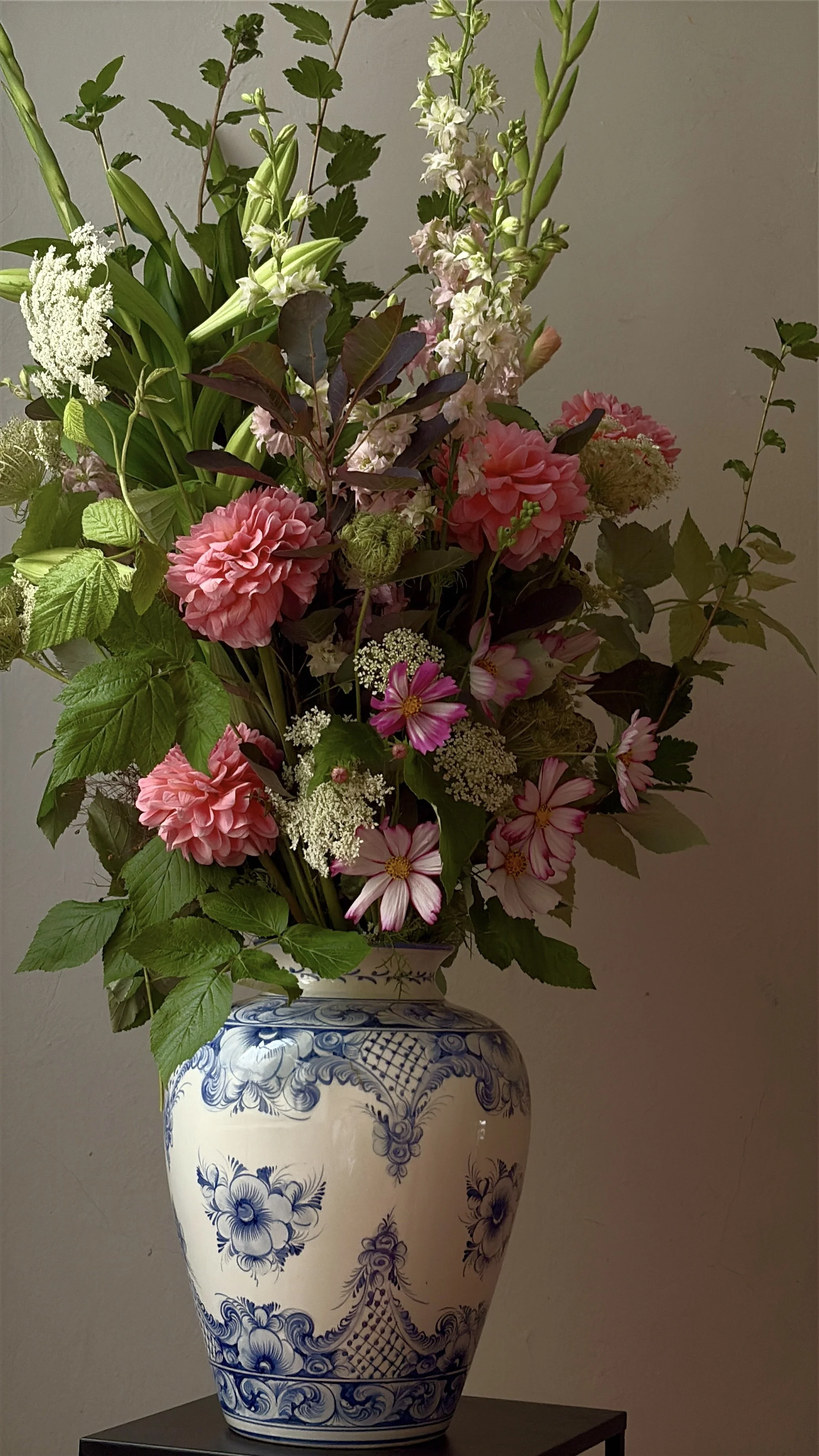 Abundant pink and white floral arrangement with dahlias, cosmos, delphiniums, greenery, and seed heads in a blue-and-white patterned ceramic vase.