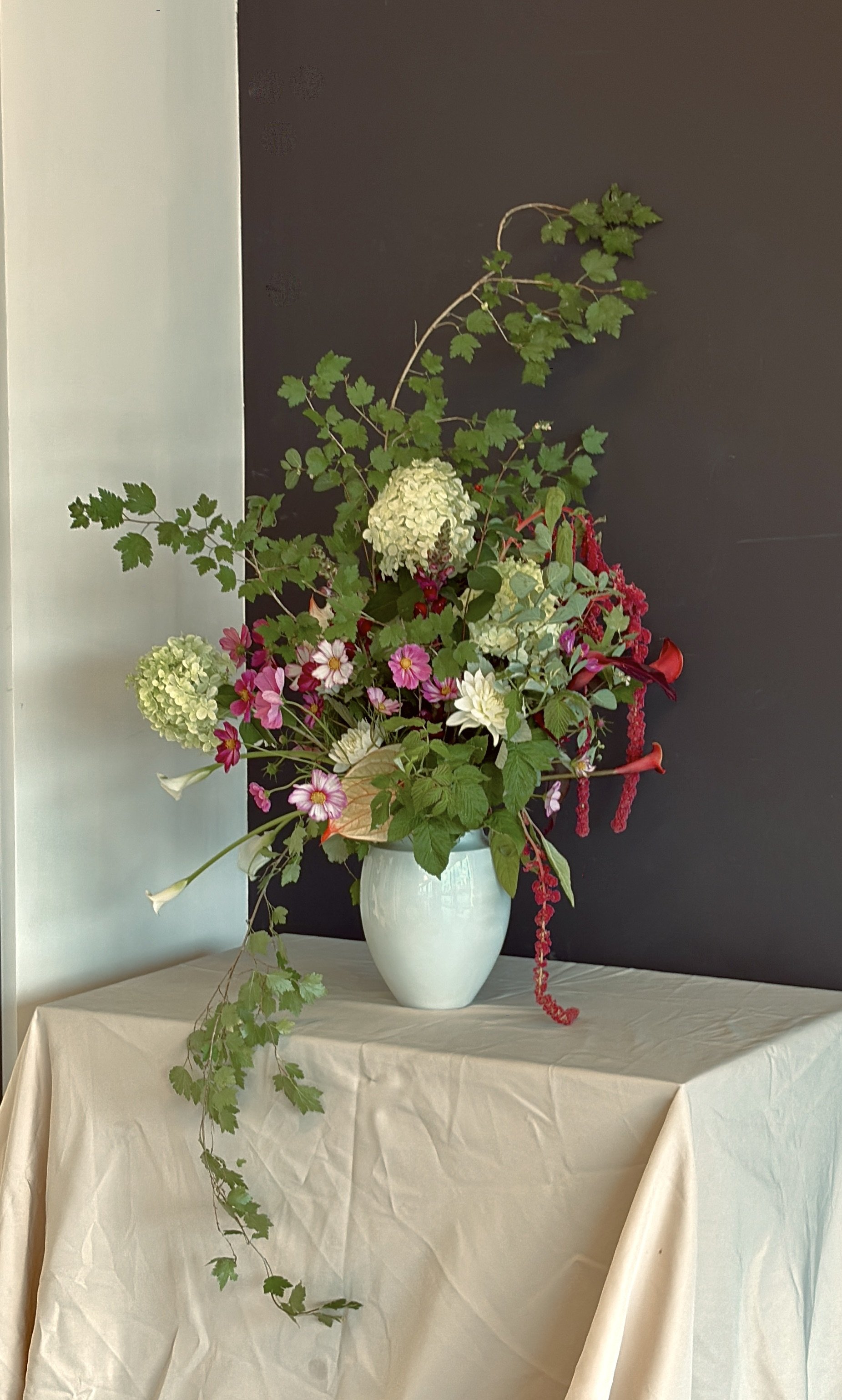Large colorful floral arrangement with hydrangeas, daisies, chrysanthemums, trailing greenery, and red flowers in a pale blue vase on a white tablecloth