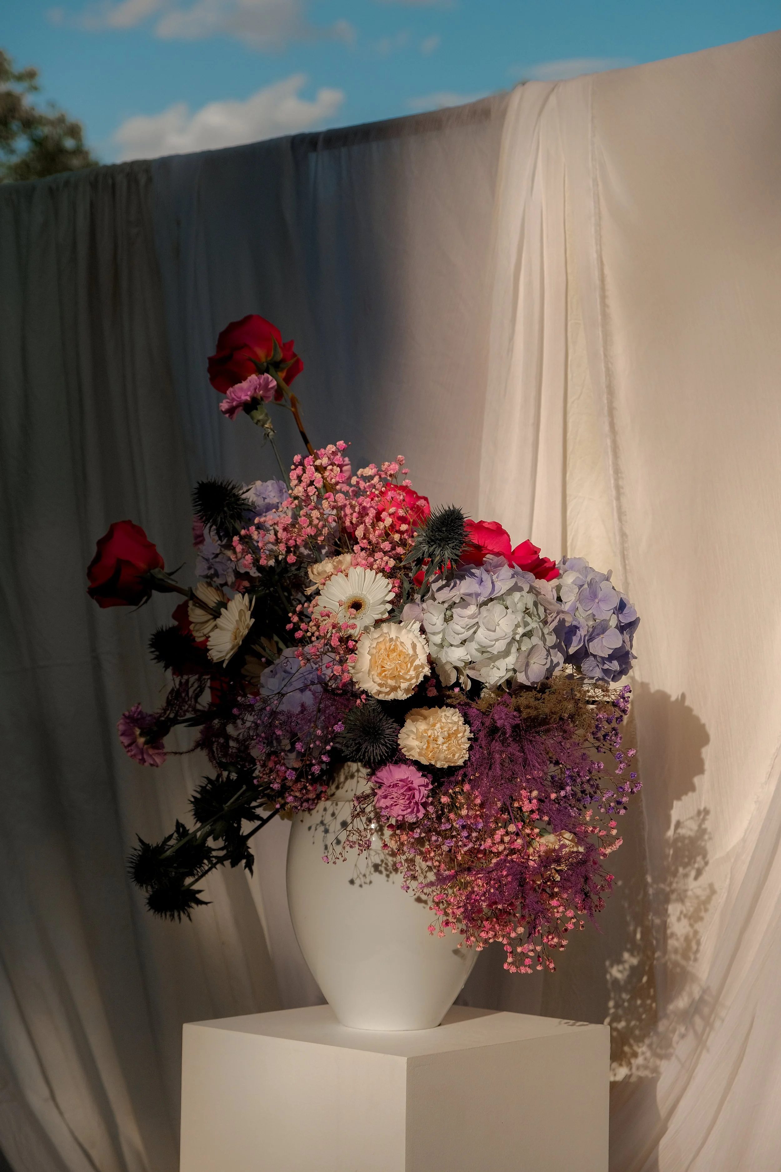 Colorful floral arrangement in a white vase on a pedestal, displayed against draped fabric with dramatic lighting.