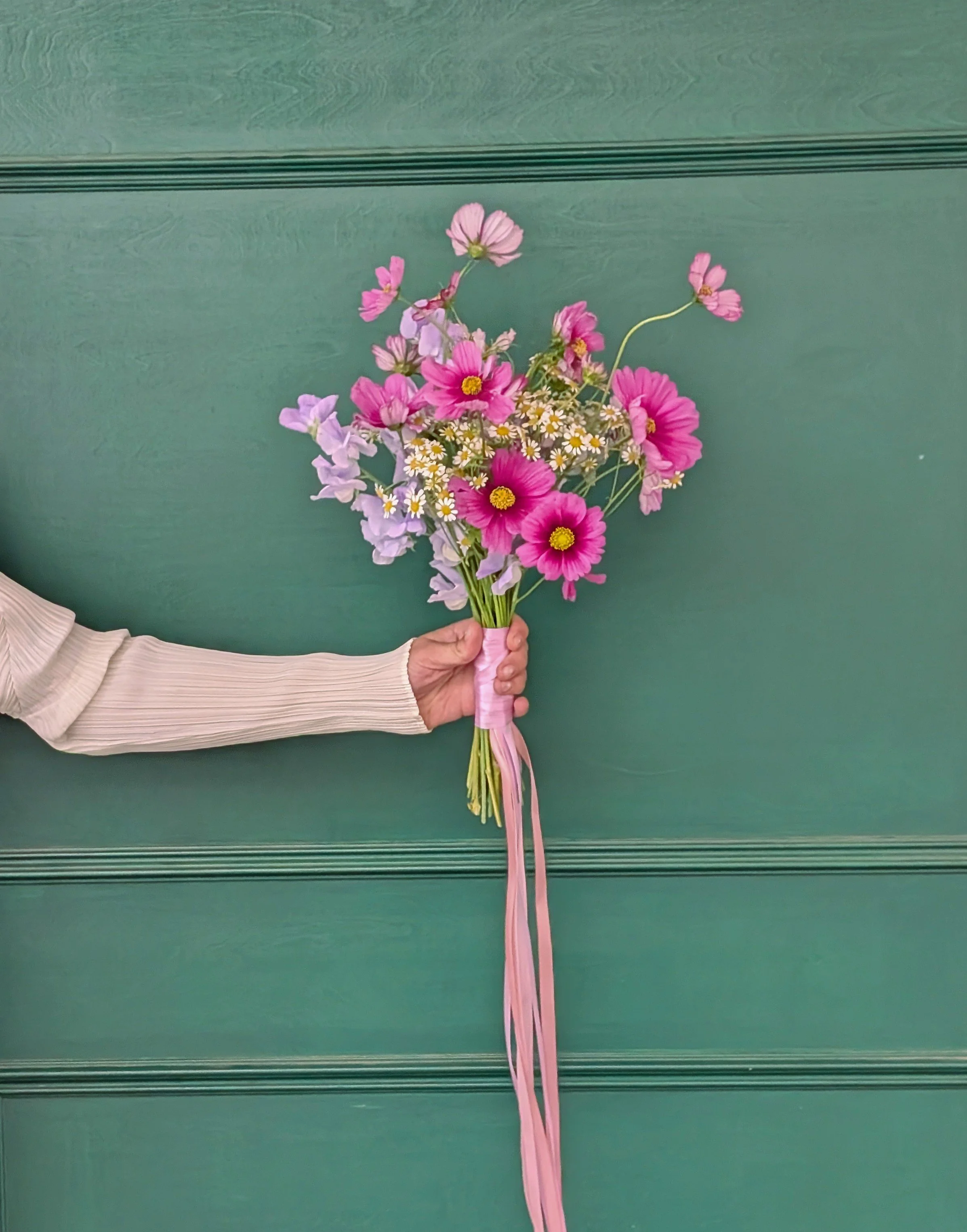 Person in a white pleated sleeve holding a pink wildflower bouquet against a teal paneled background