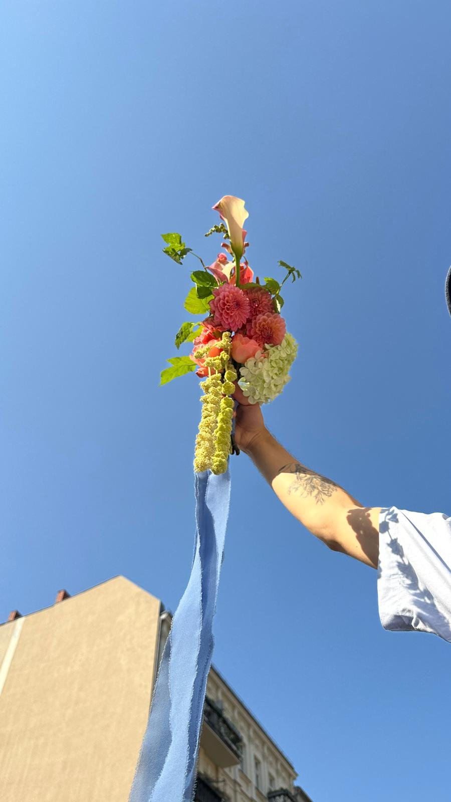 Hand holding a colorful floral arrangement with trailing blue ribbons against a clear blue sky
