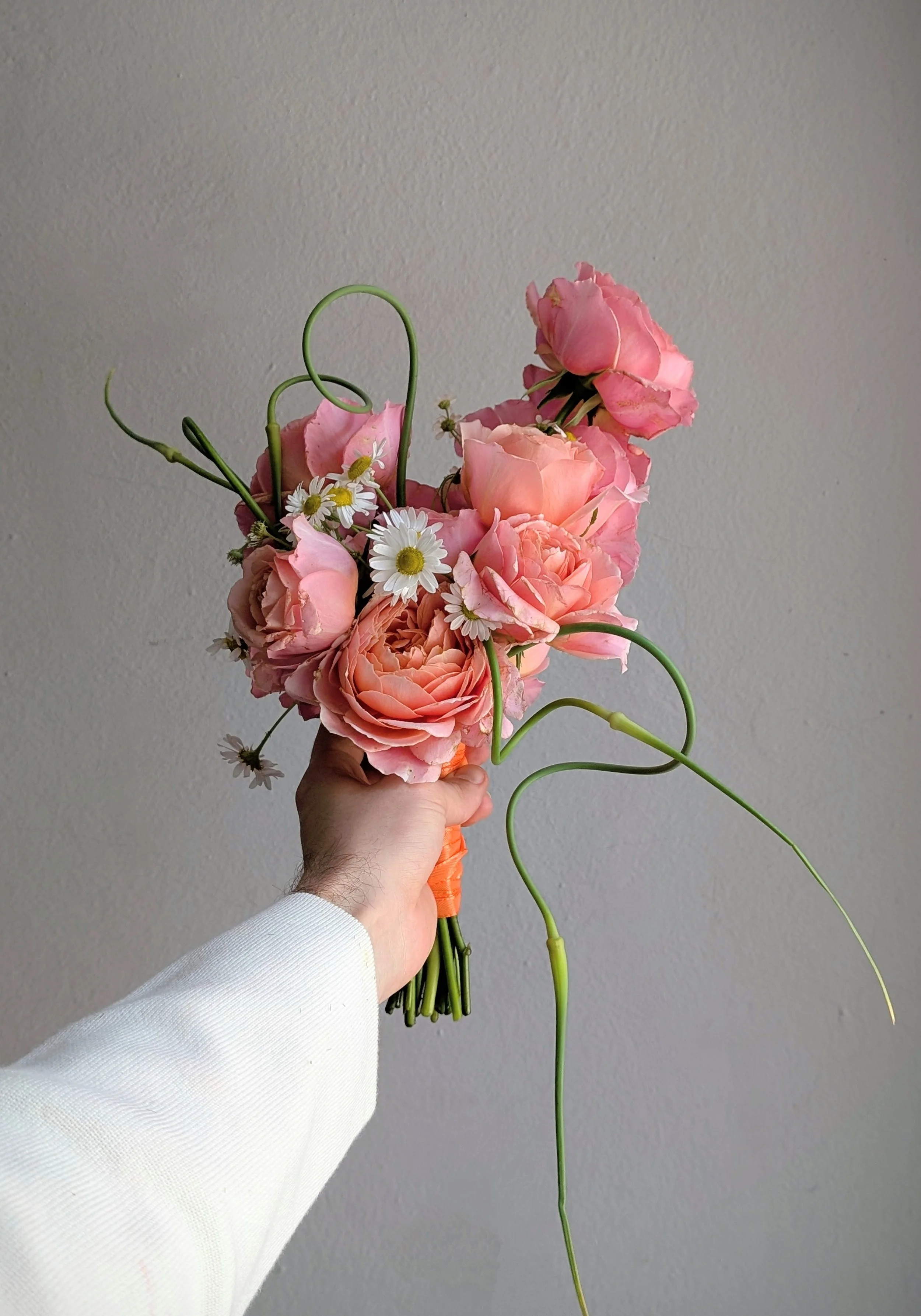 Hand holding a pink rose bouquet with white daisies and curling green stems against a gray wall