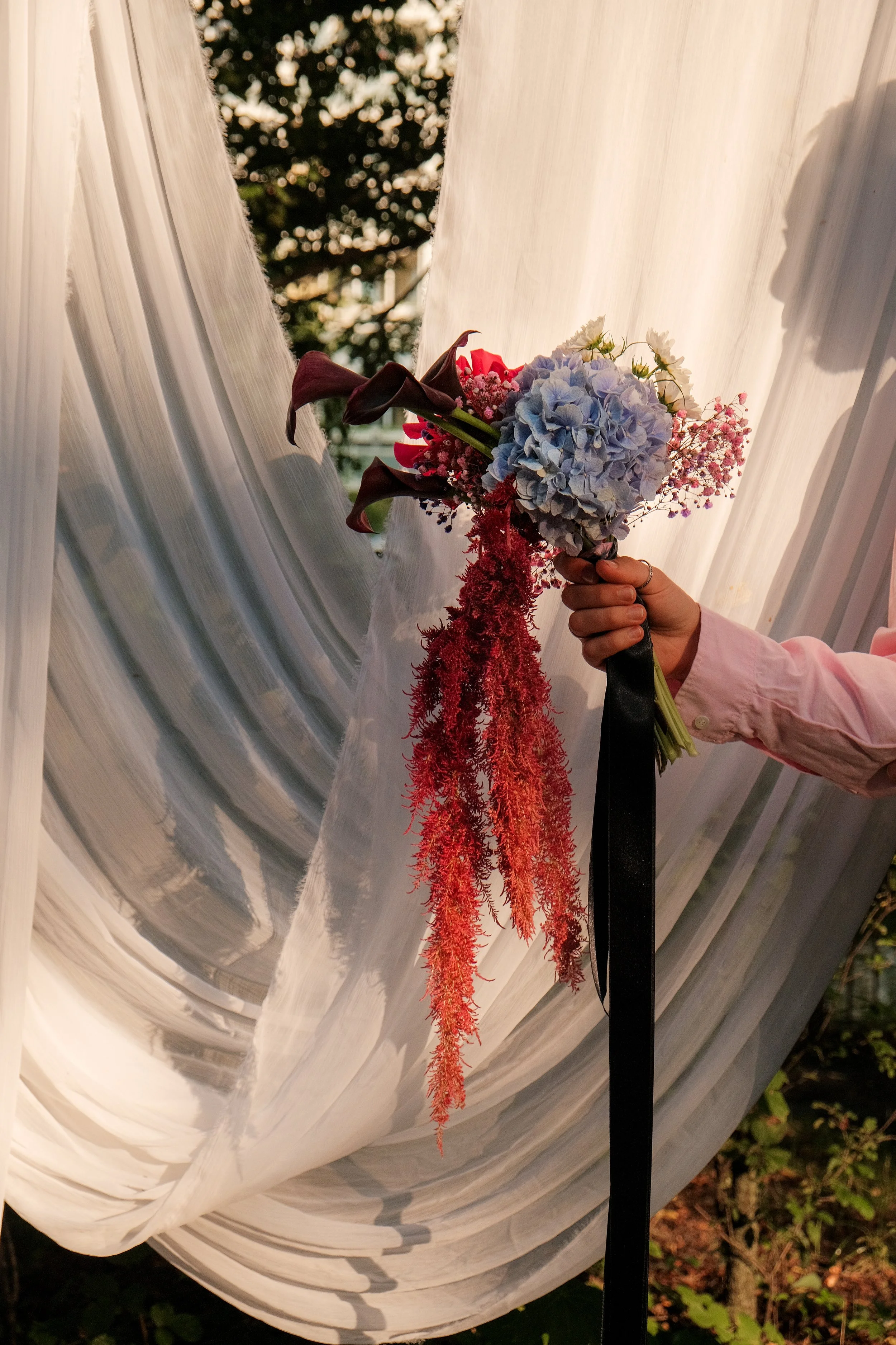 Person in a pink shirt holding a colorful bouquet of hydrangeas and dried flowers in front of flowing white curtains